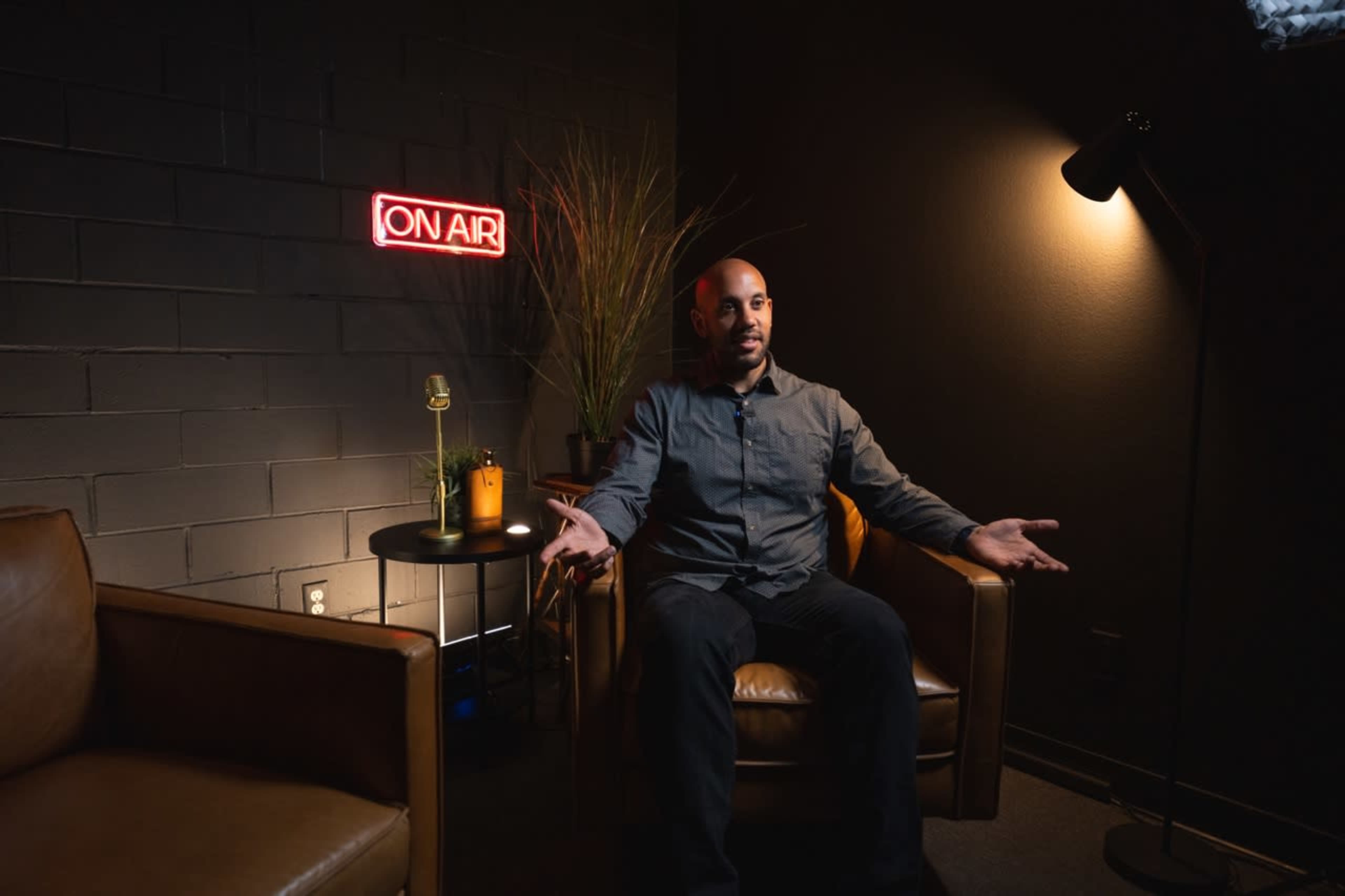 A man sits in a brown leather chair in a dimly lit room with an "ON AIR" sign and a table with a microphone and a candle.