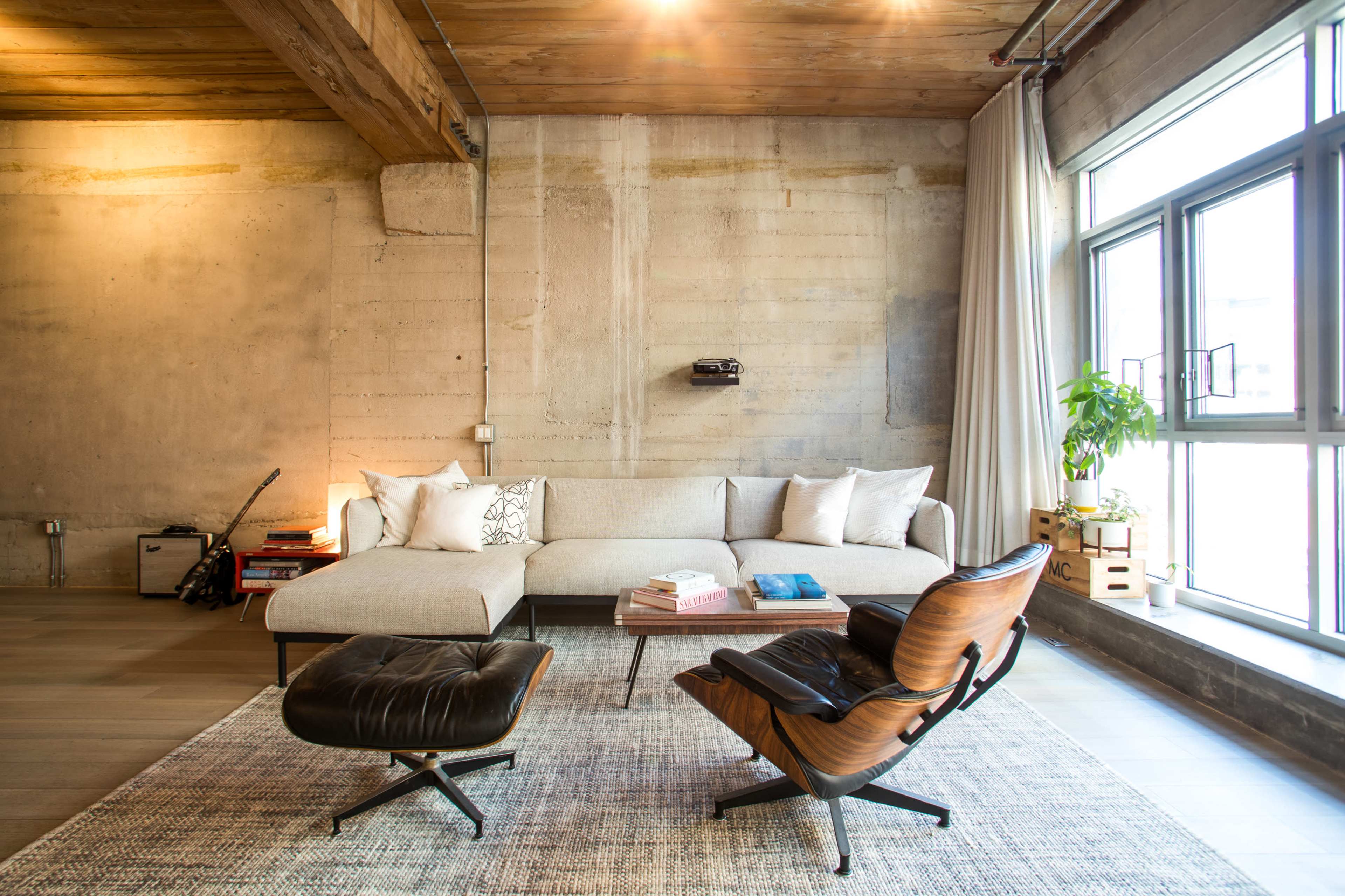 The image shows a modern living room with a sectional sofa, a wooden chair, a coffee table, and large windows letting in natural light.