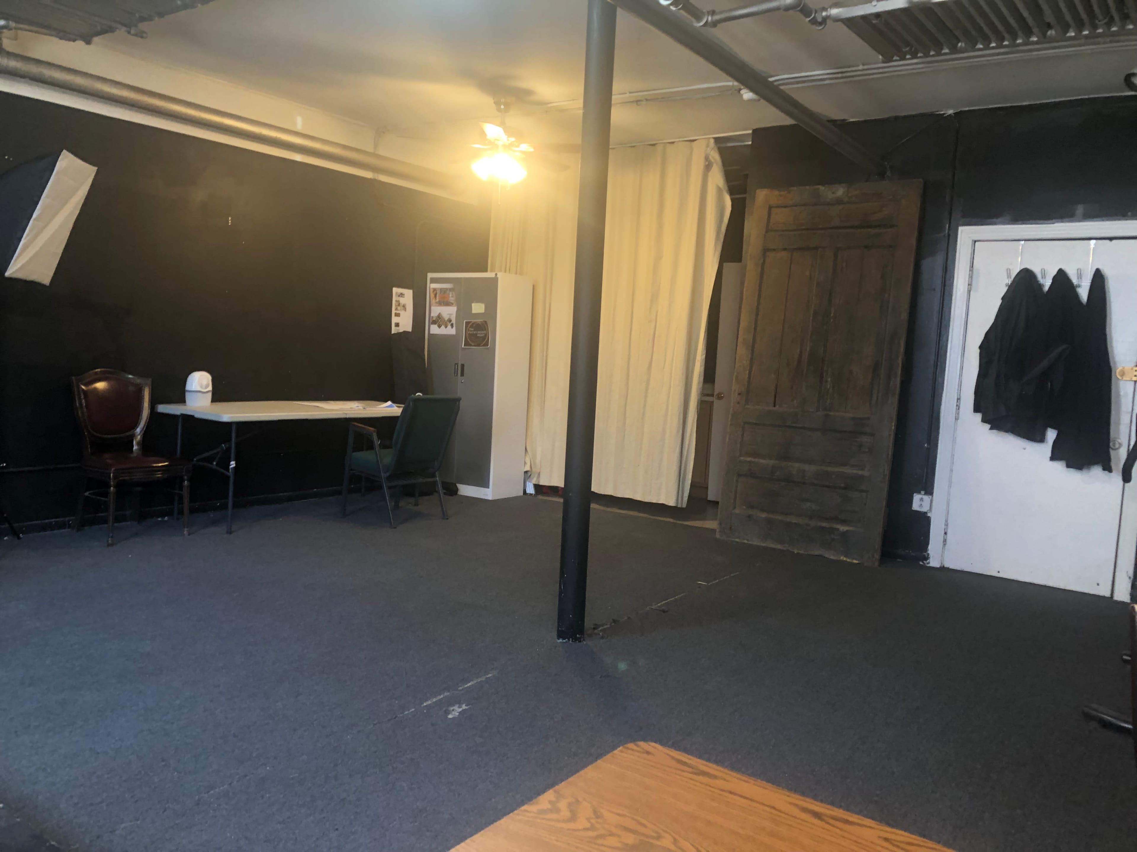 An empty room with a dark wall, a table and two chairs, a white cabinet, a large wooden door, and a curtain partition.