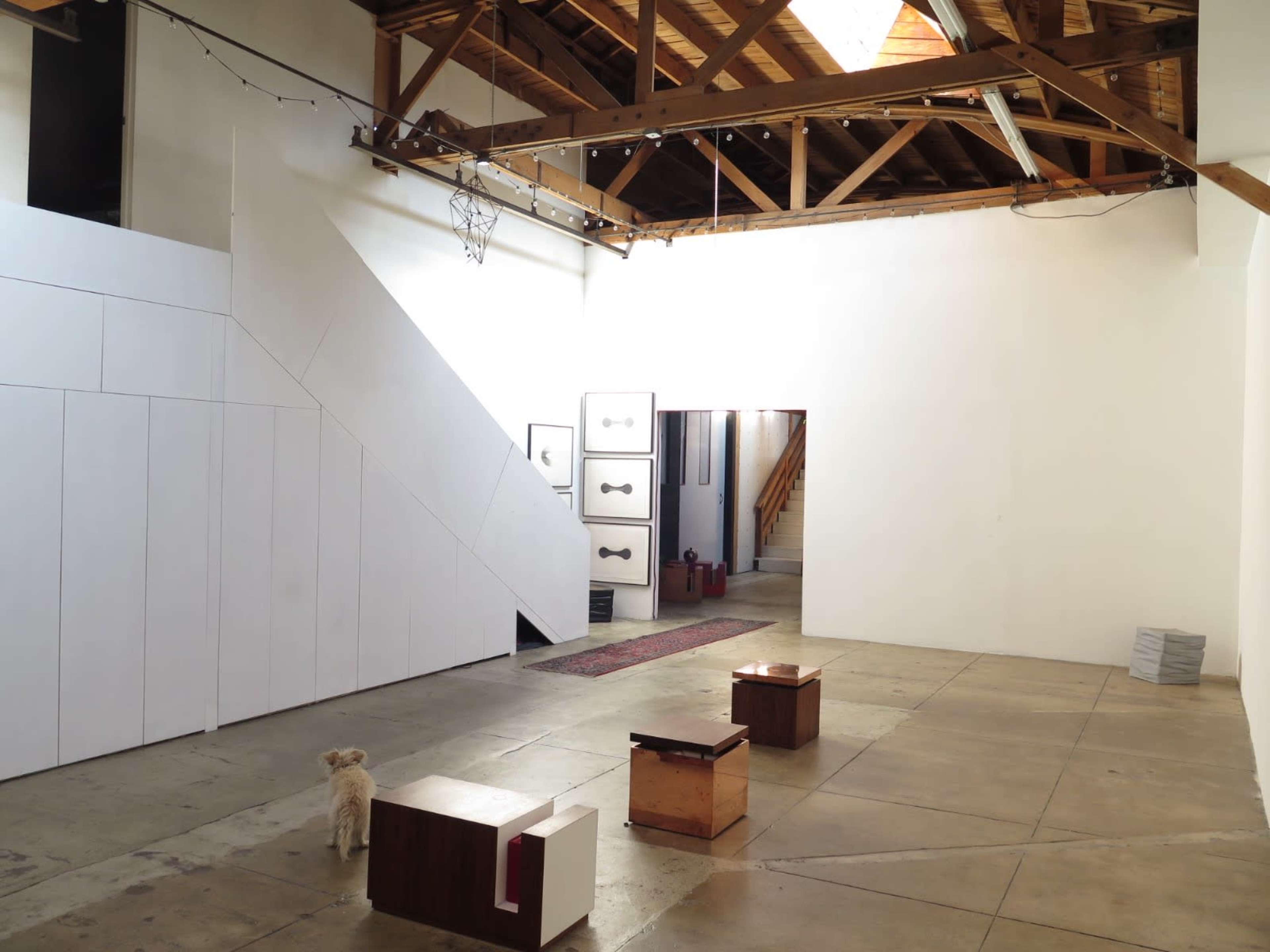 An empty gallery space features a polished concrete floor, exposed wooden beams, and a small dog standing near abstract wooden sculptures.
