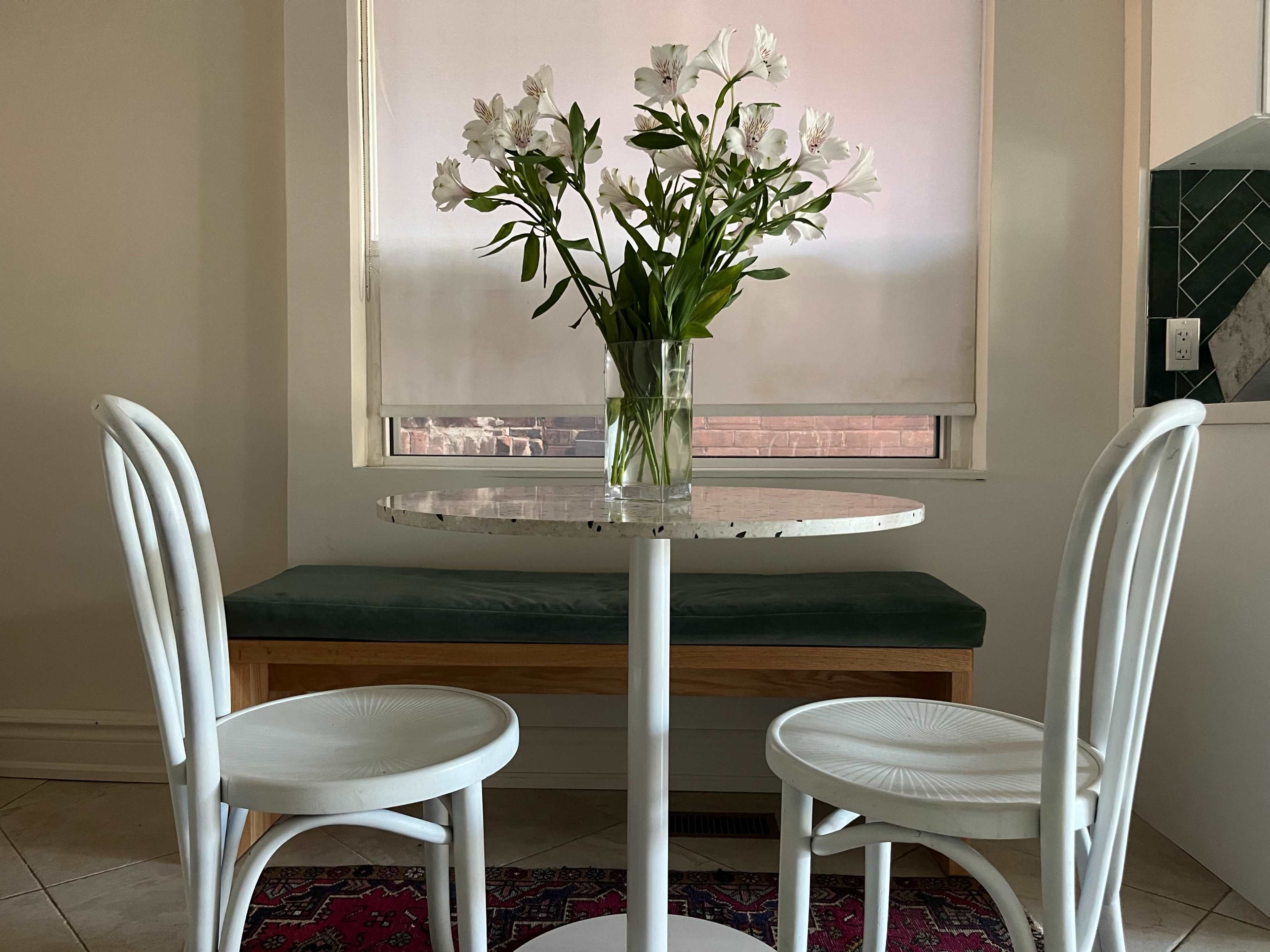 A small, round table with two white chairs is arranged in a bright room, featuring a vase of white flowers on the table and a green cushion bench in the background.