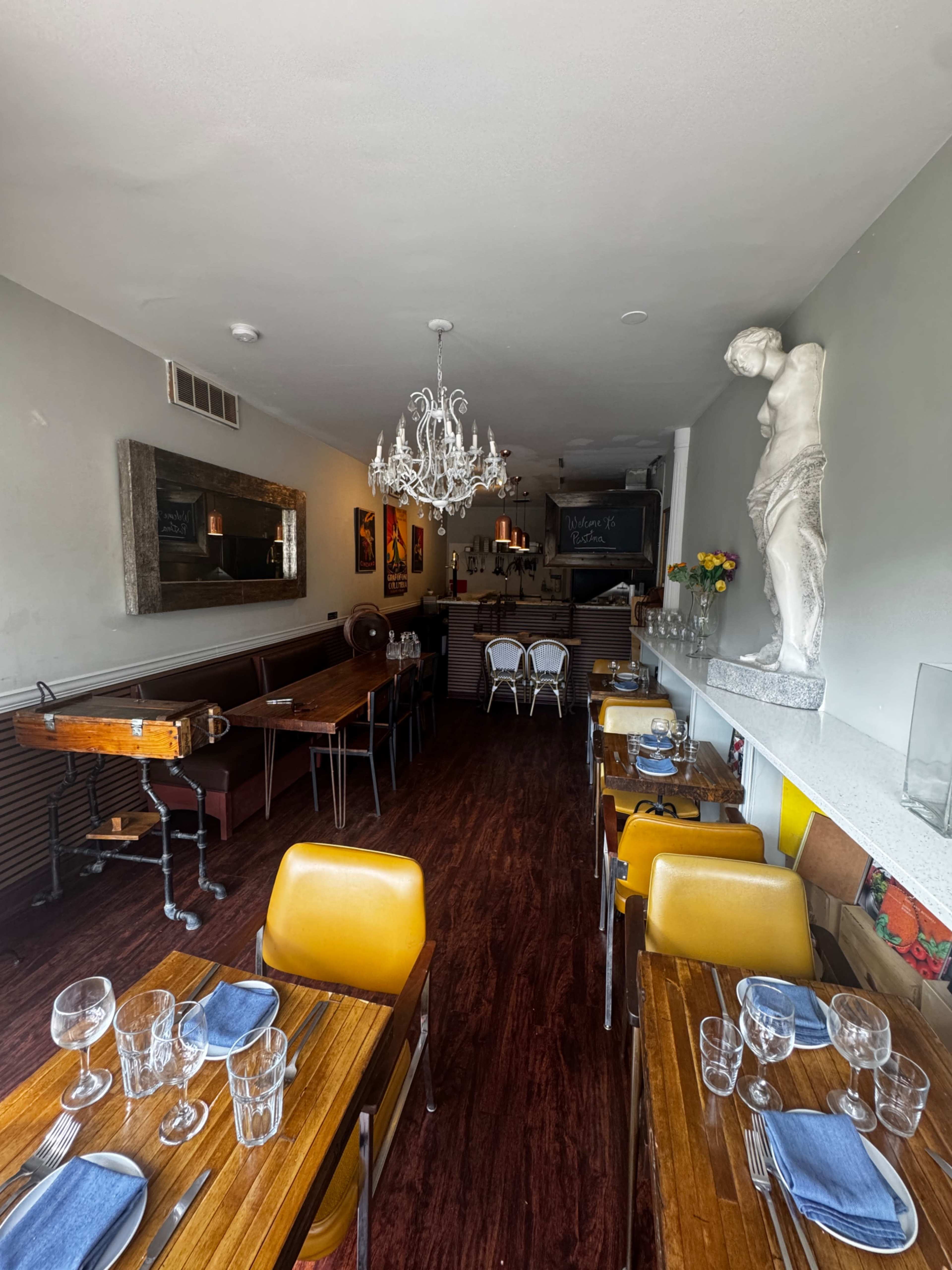 The image shows a narrow restaurant interior with wooden tables set with blue napkins and glasses, a chandelier overhead, and a statue displayed against a wall.