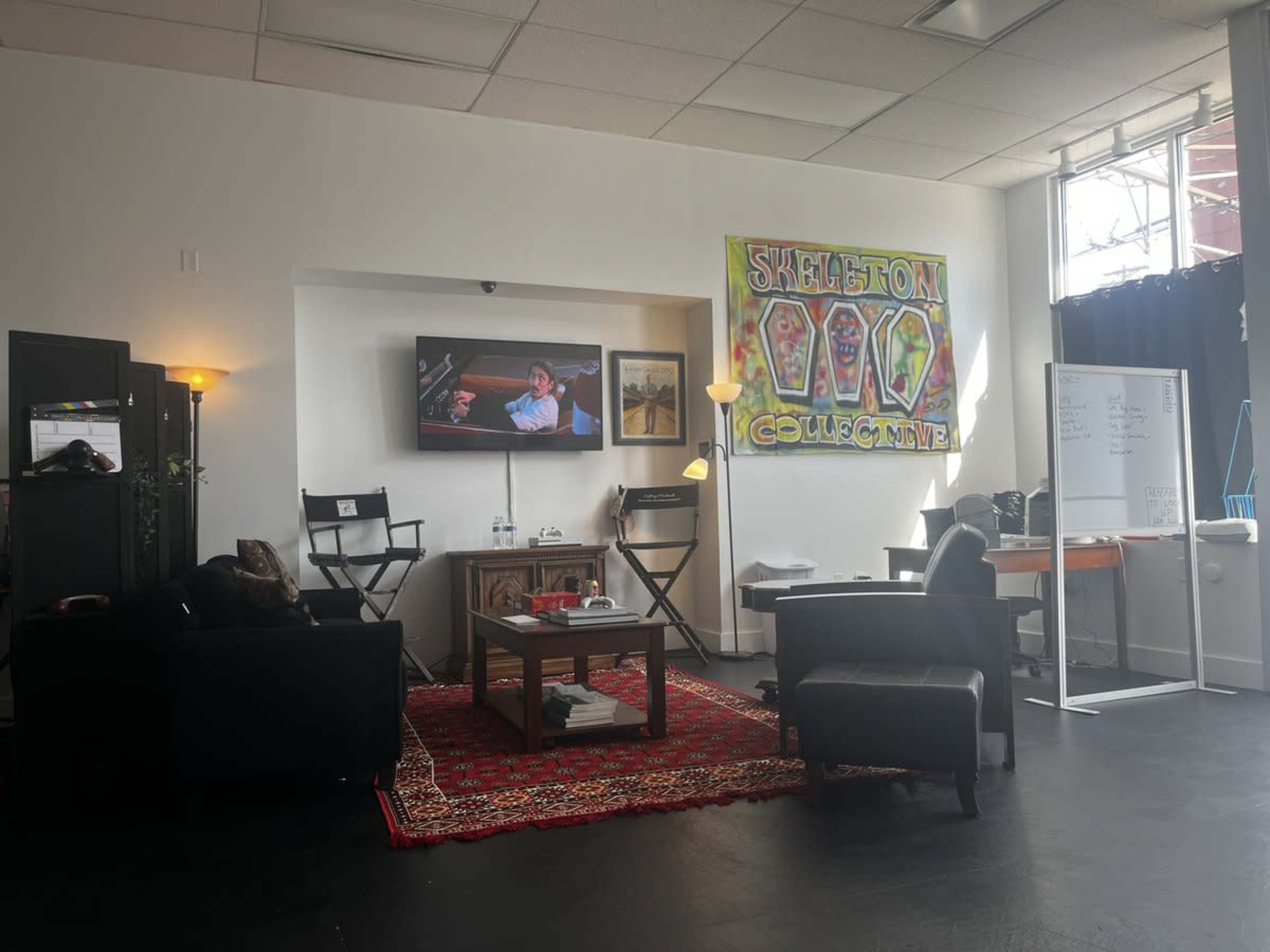 The image shows a cozy studio space with black chairs, a television displaying a scene, a small table, a red rug, and colorful artwork on the walls.