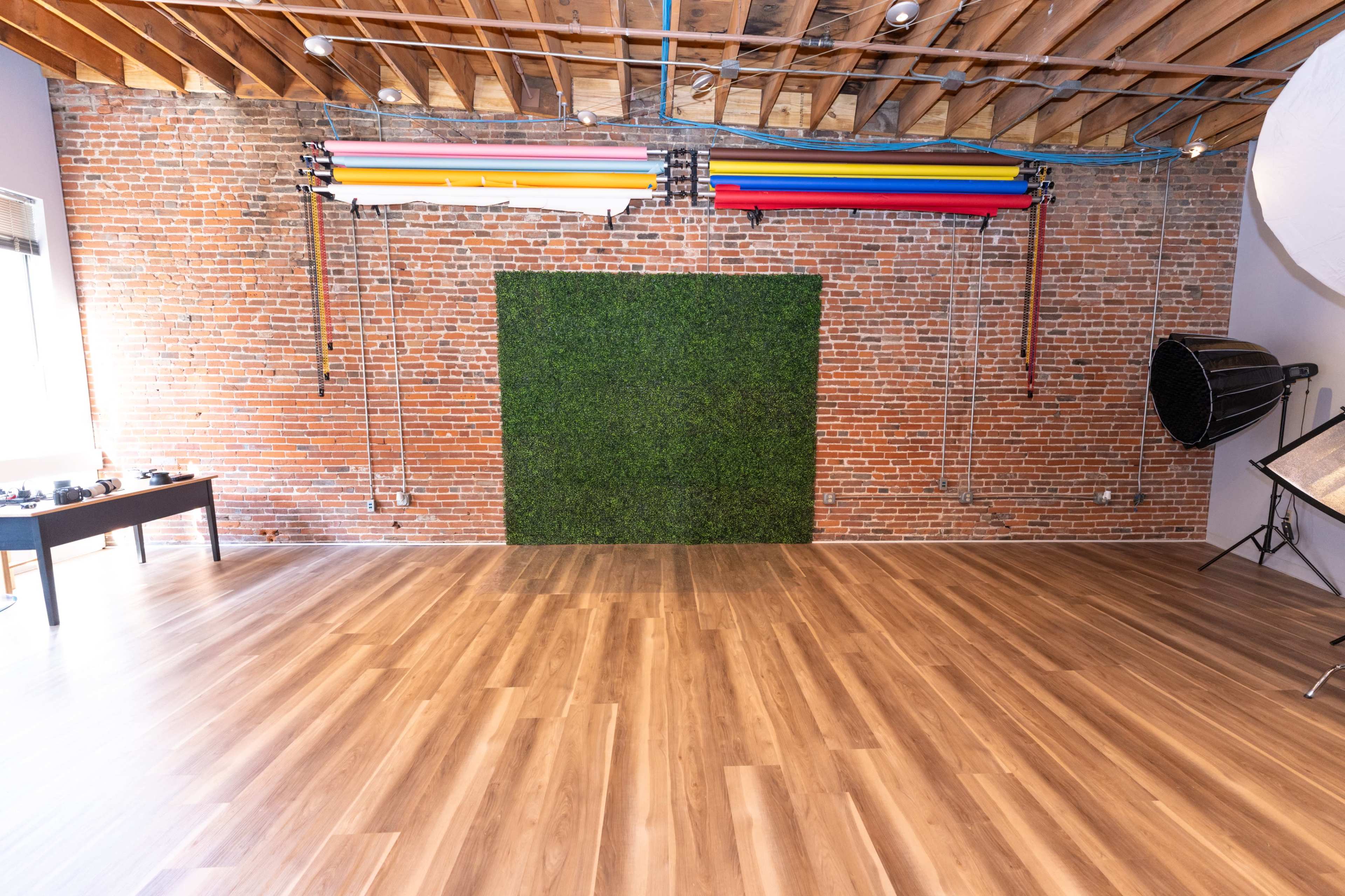 The image shows a spacious, empty studio featuring a brick wall with a green grass backdrop and colorful backdrops mounted above, alongside a wooden floor.