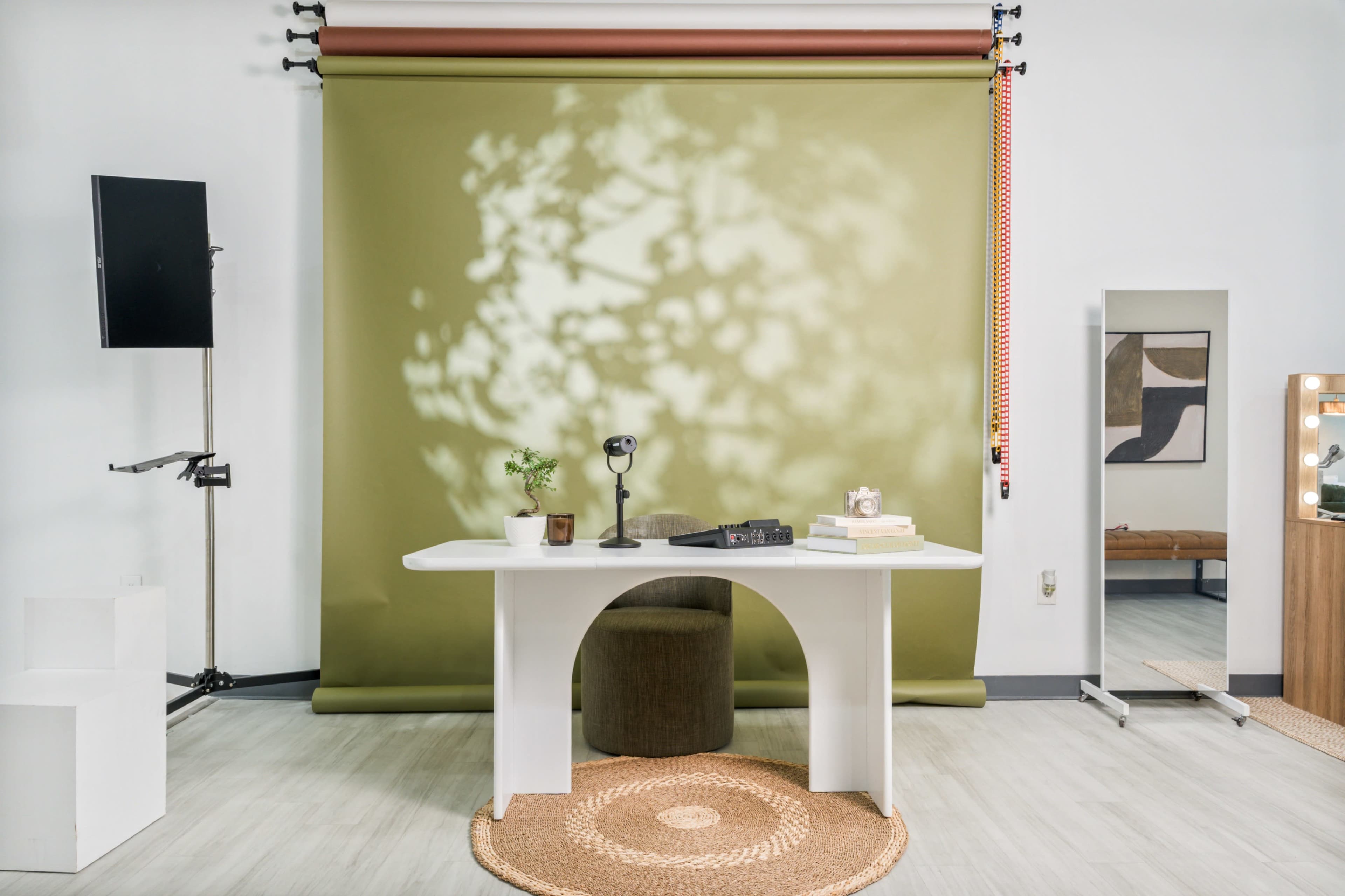 A modern studio space features a white table with a microphone and a plant, set against a green backdrop with shadows and a round mirror nearby.