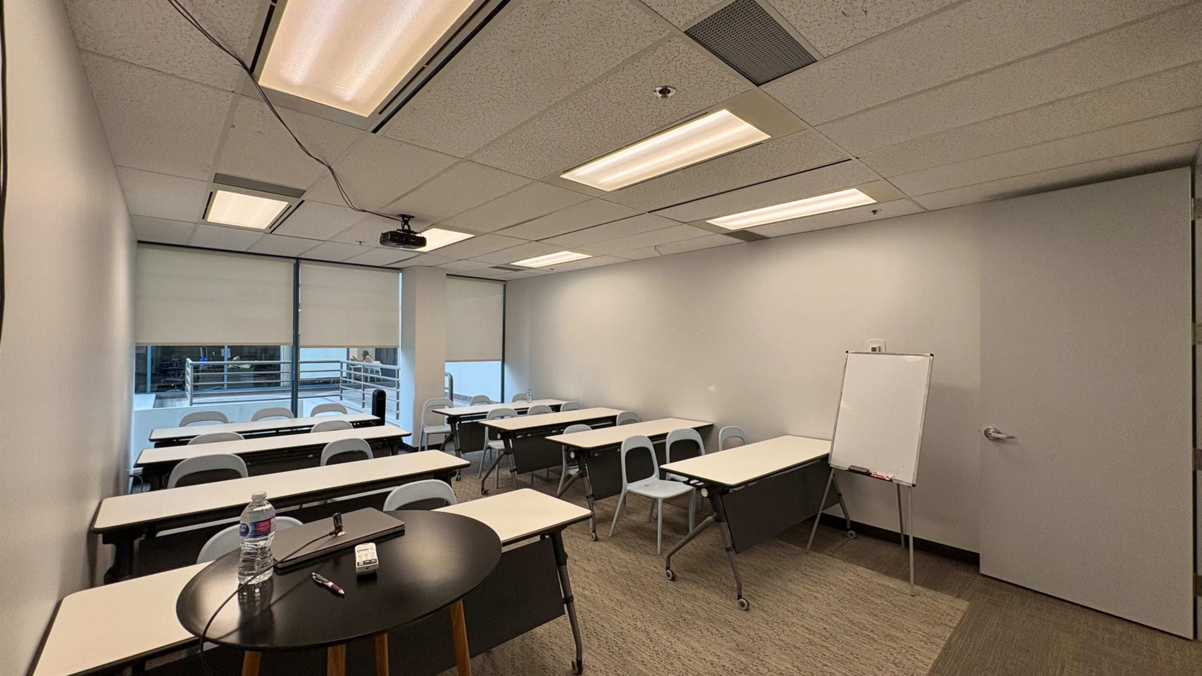 The image shows a classroom with several rows of tables and chairs, a whiteboard, and a projector mounted on the ceiling.