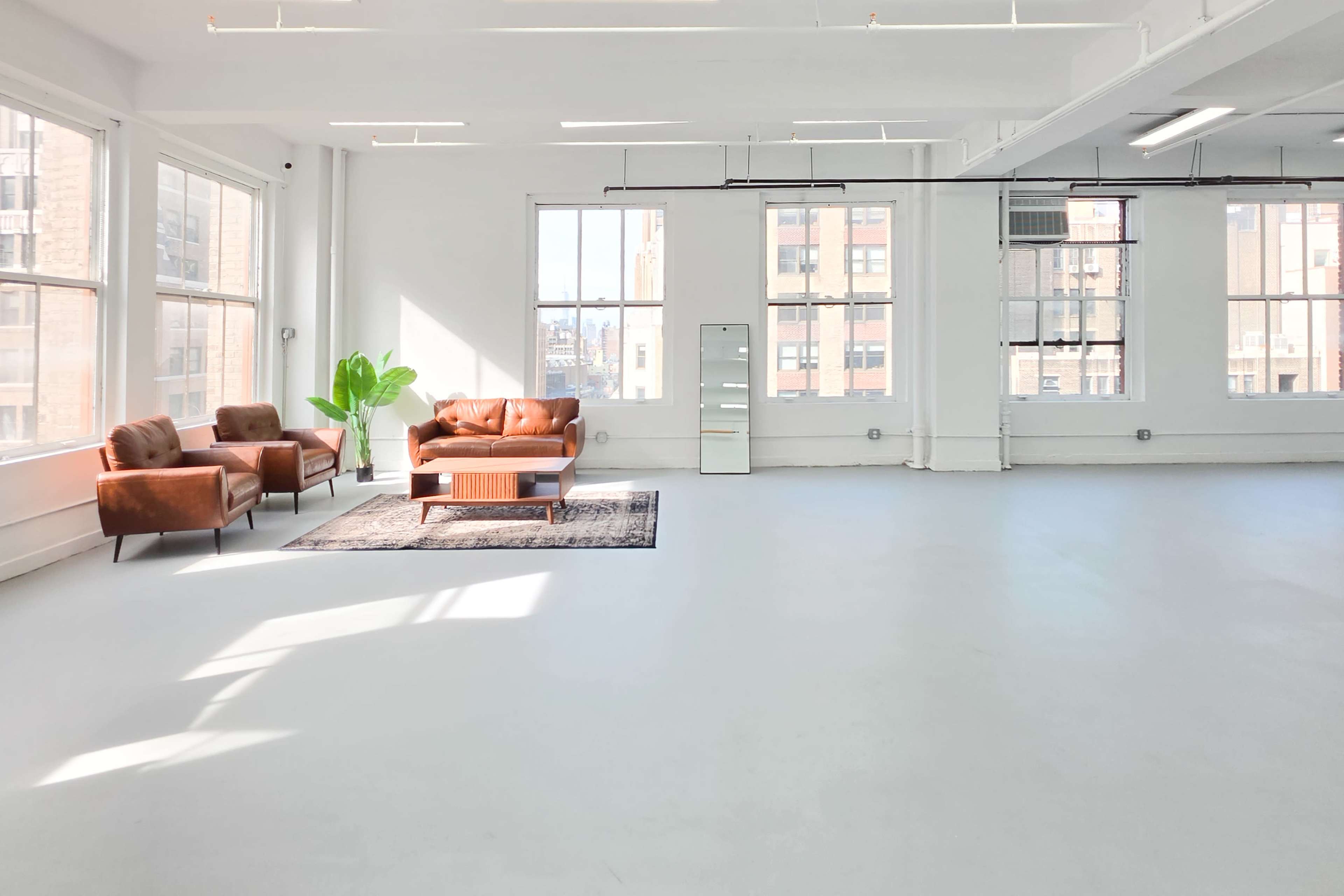 The image shows a bright, spacious room featuring a brown leather sofa and chairs, a wooden coffee table, and large windows allowing ample natural light.
