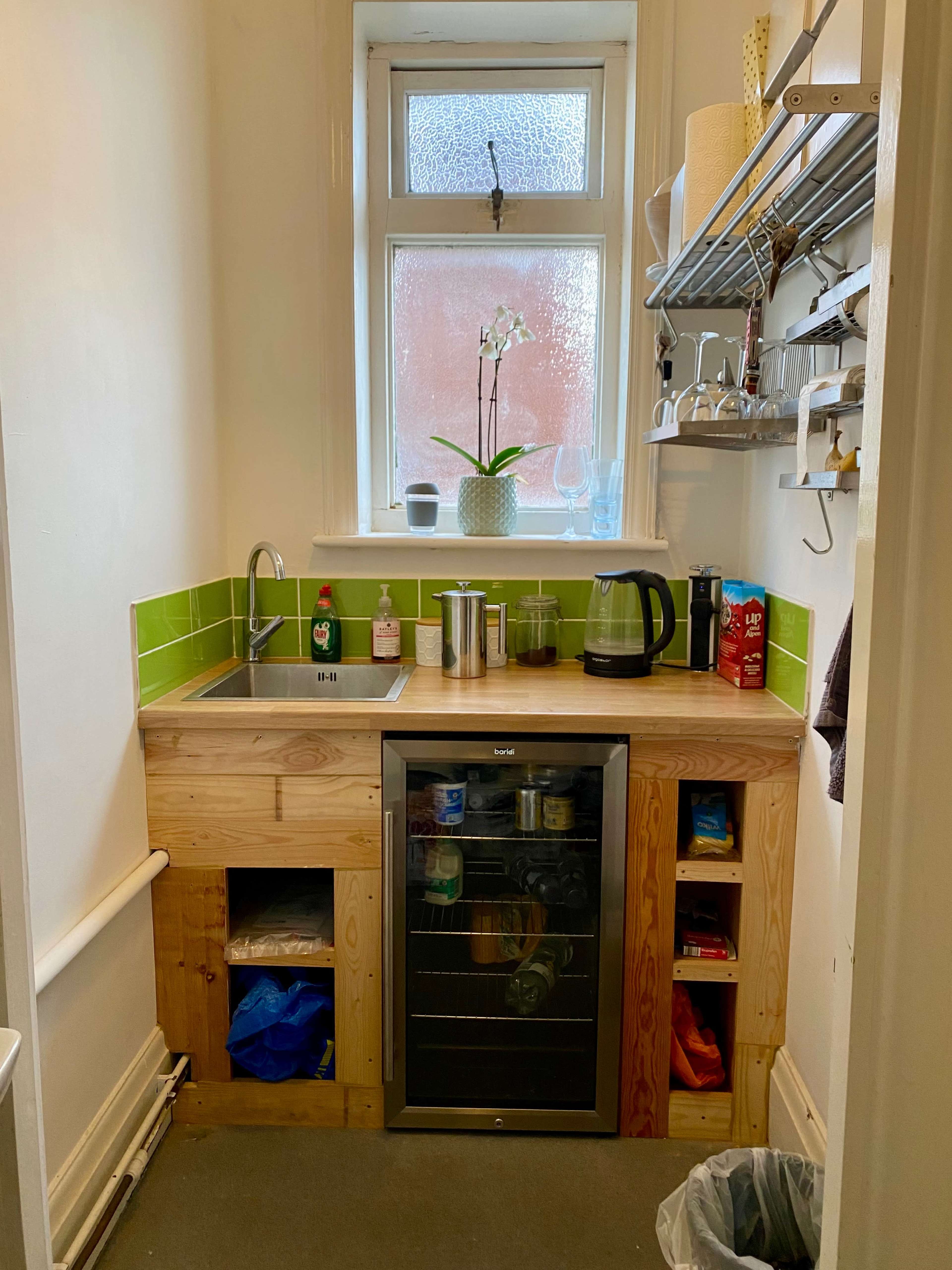 A small kitchen area featuring a wooden cabinet with a sink, a kettle, and a compact refrigerator, along with green tiled backsplash and a window above with a potted plant.