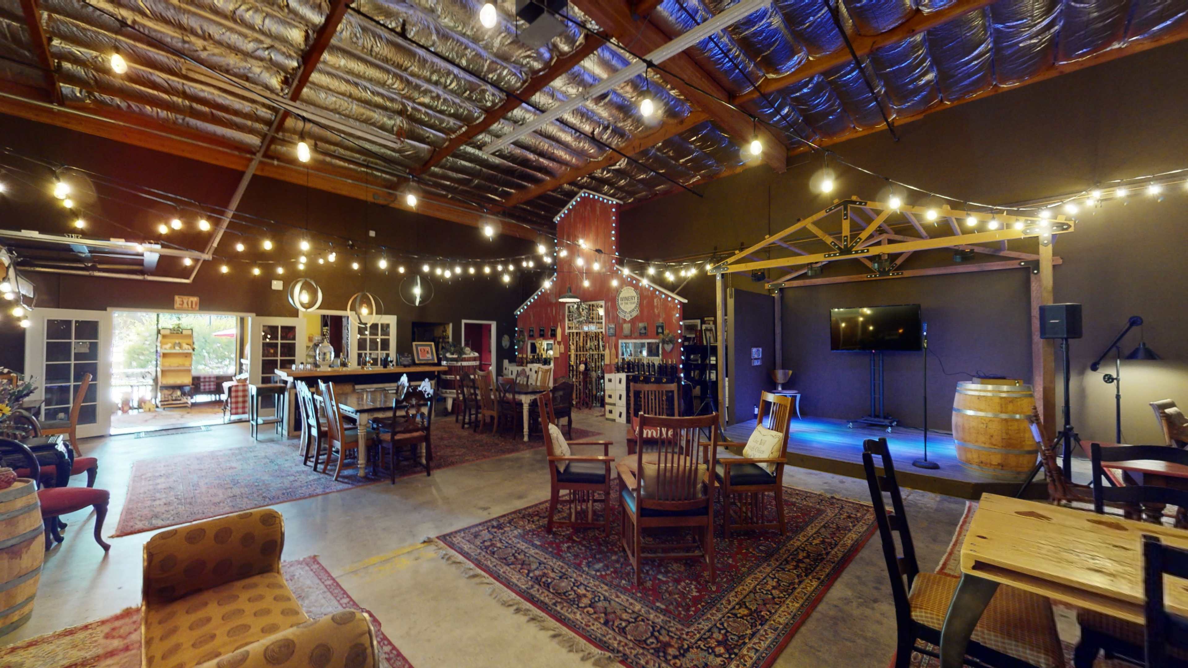 An indoor space with wooden furniture, string lights, and a stage area, resembling a rustic gathering venue.