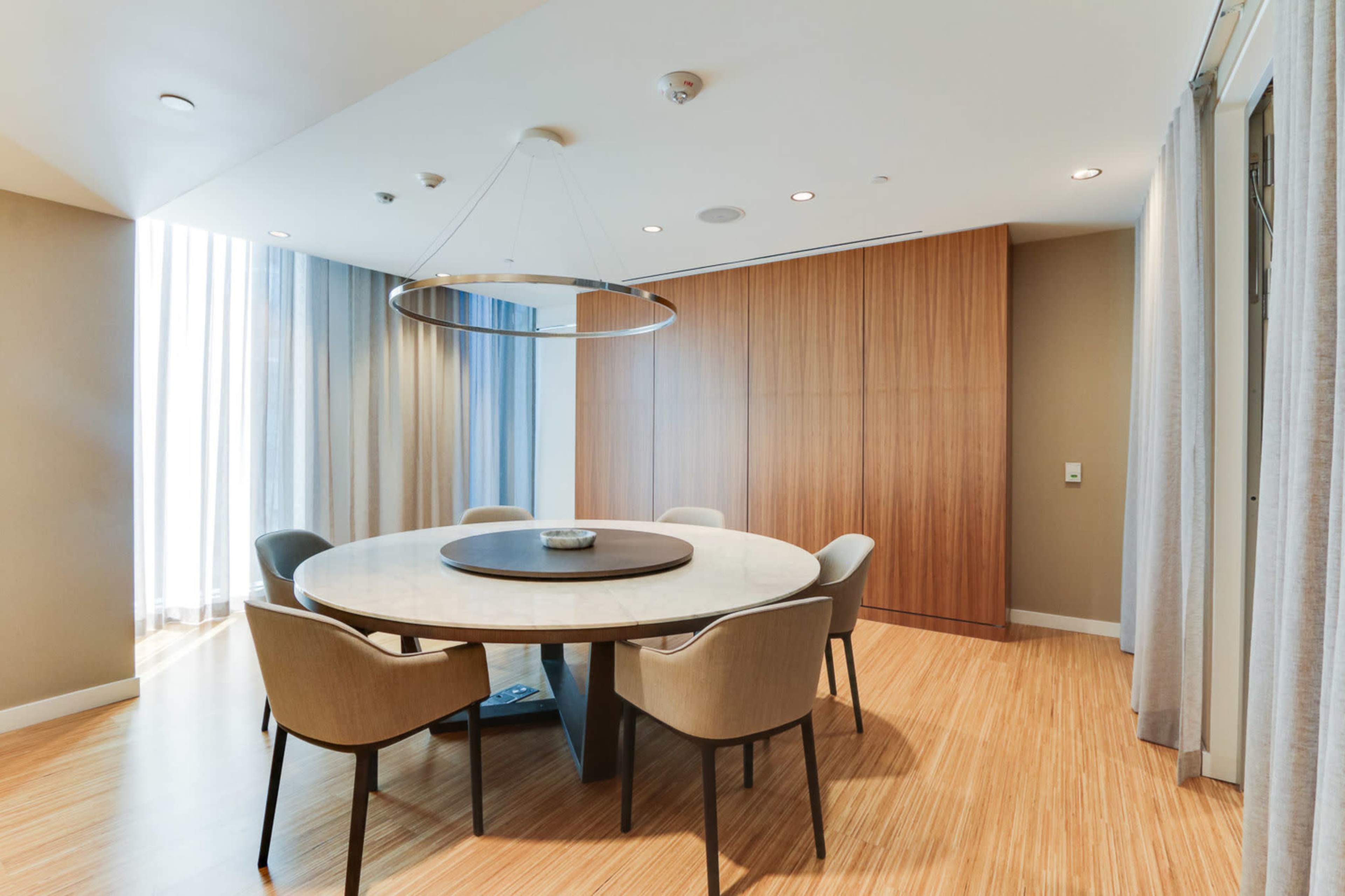 A round dining table with six chairs is positioned in a well-lit room with wooden flooring and large windows.