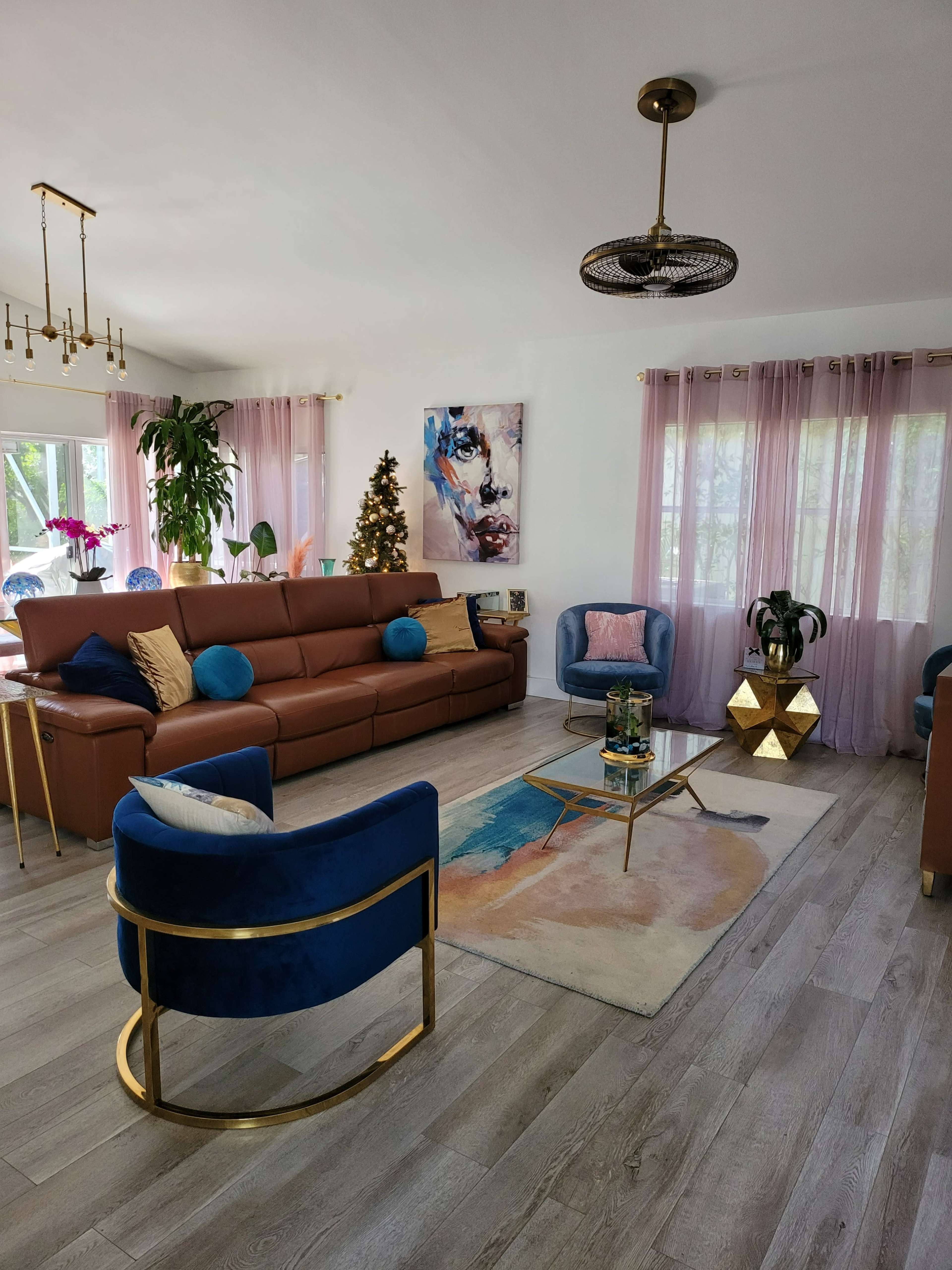 A spacious living room featuring a brown leather sofa with blue and gold cushions, two blue accent chairs, a coffee table, and a Christmas tree decorated in the corner.