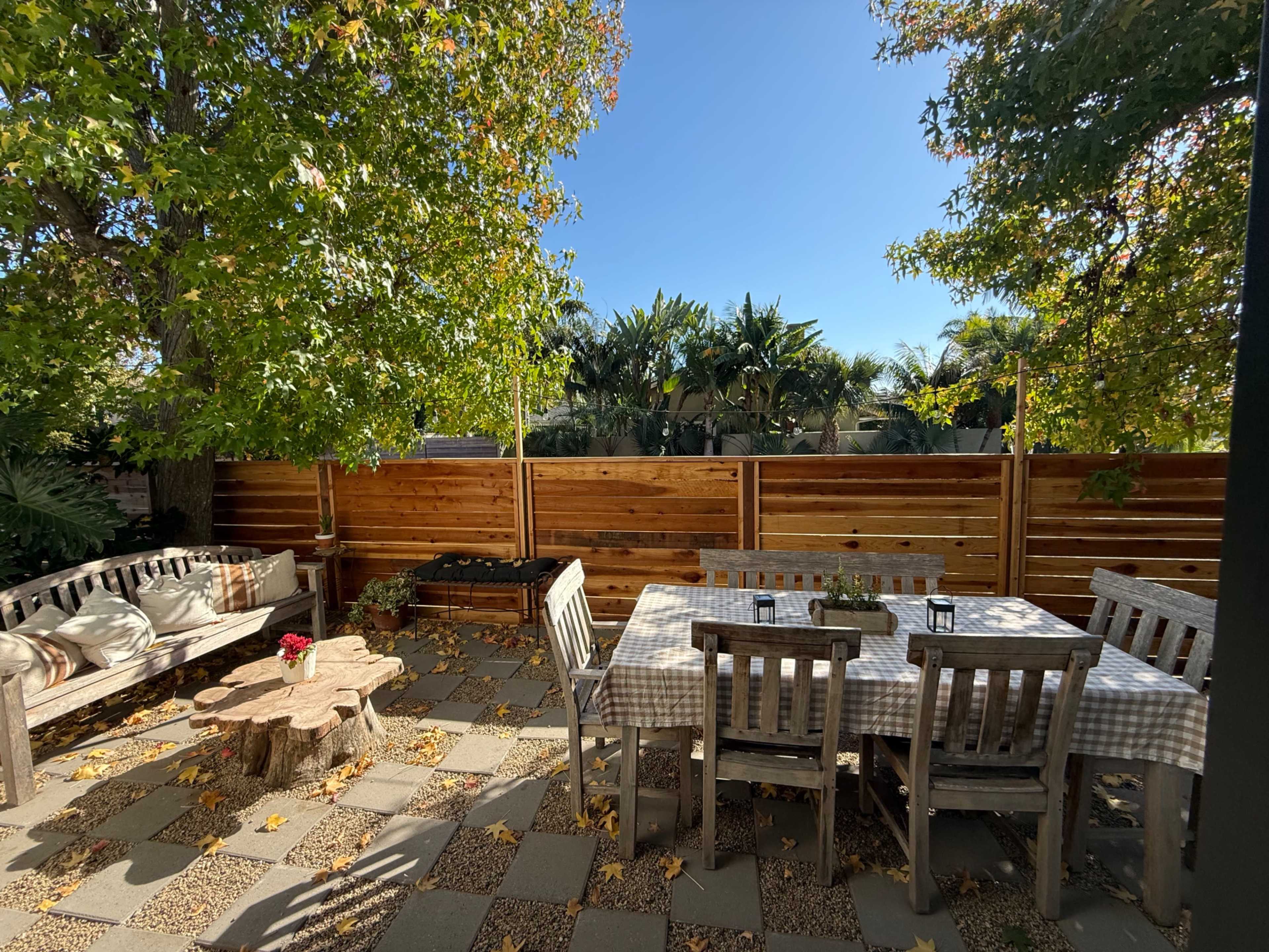 A backyard patio features a wooden dining set, a coffee table made from a tree stump, and a sofa surrounded by trees and a wooden fence under clear blue skies.