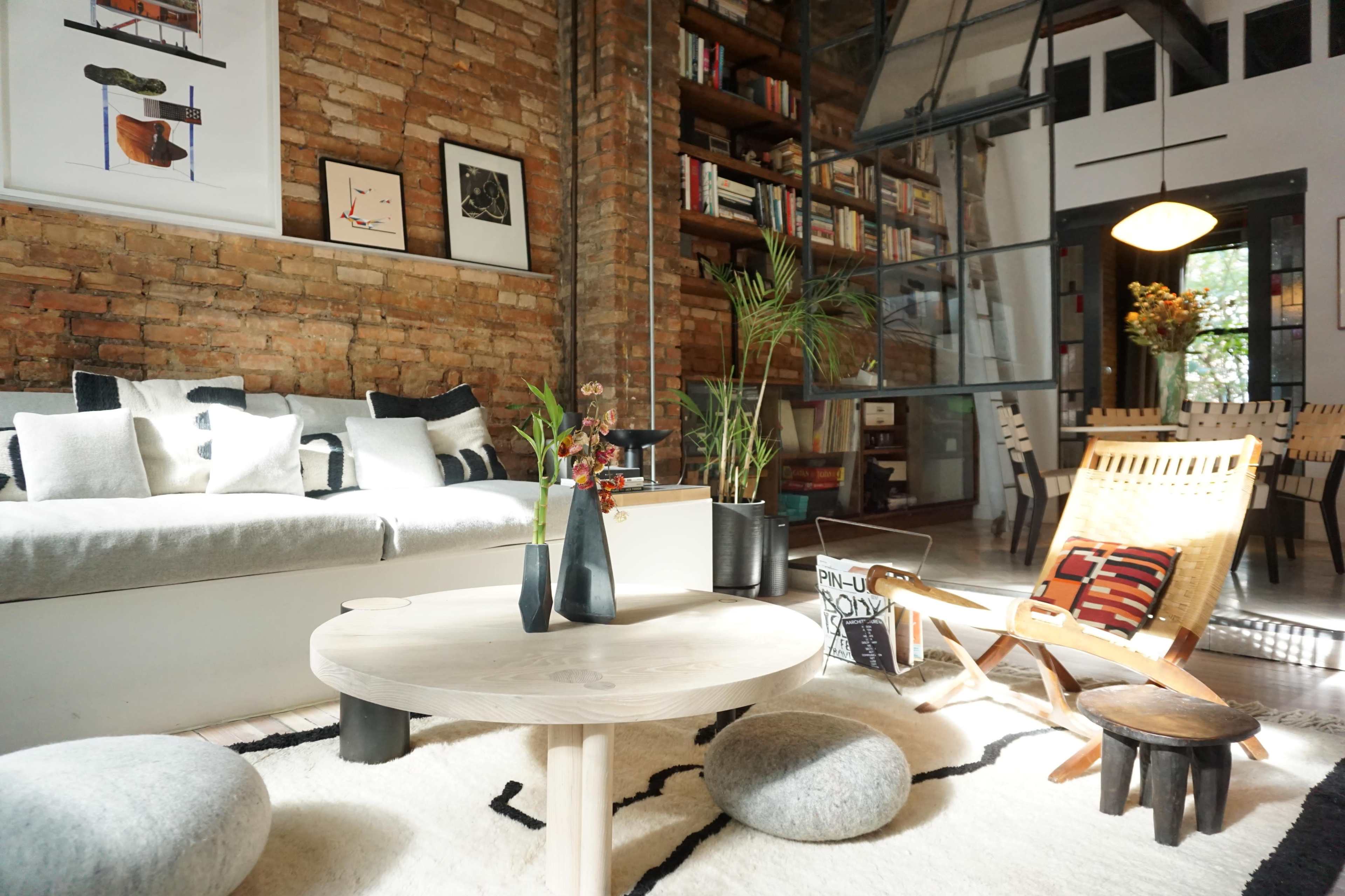 A stylish living room featuring a gray sofa, a round coffee table, and a wooden dining area against a backdrop of exposed brick walls and bookshelves.