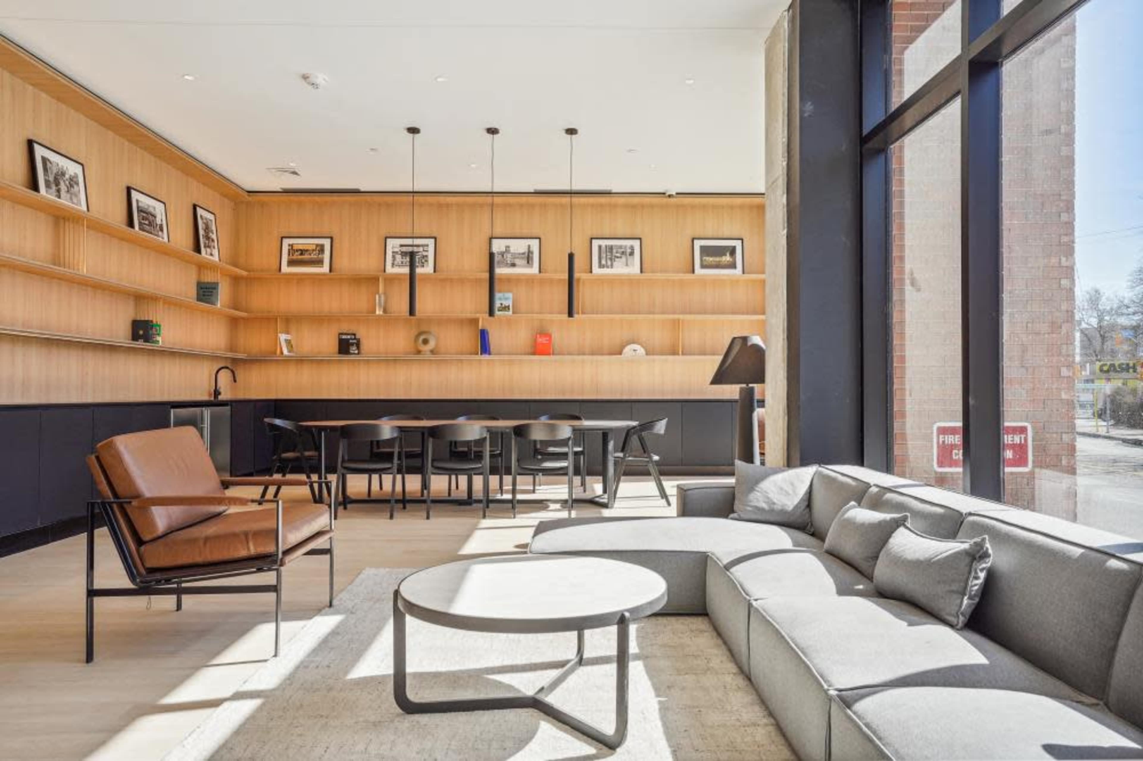A modern interior space featuring a cozy seating area with a curved gray sofa and a brown armchair, along with a wooden shelf displaying framed pictures and a dining area in the background.