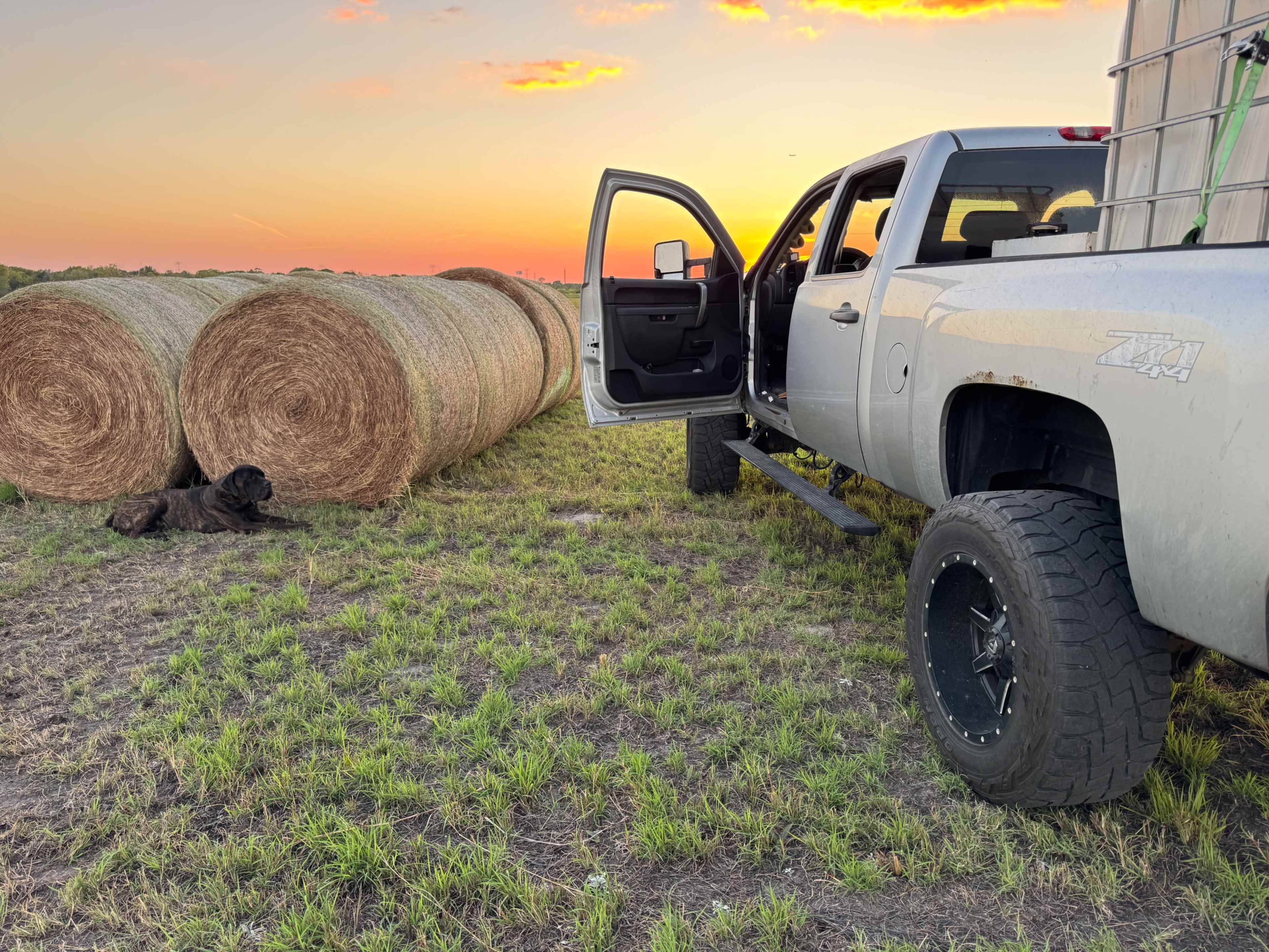A silver pickup truck is parked beside large hay bales under a colorful sunset, with a dog lying on the ground nearby.