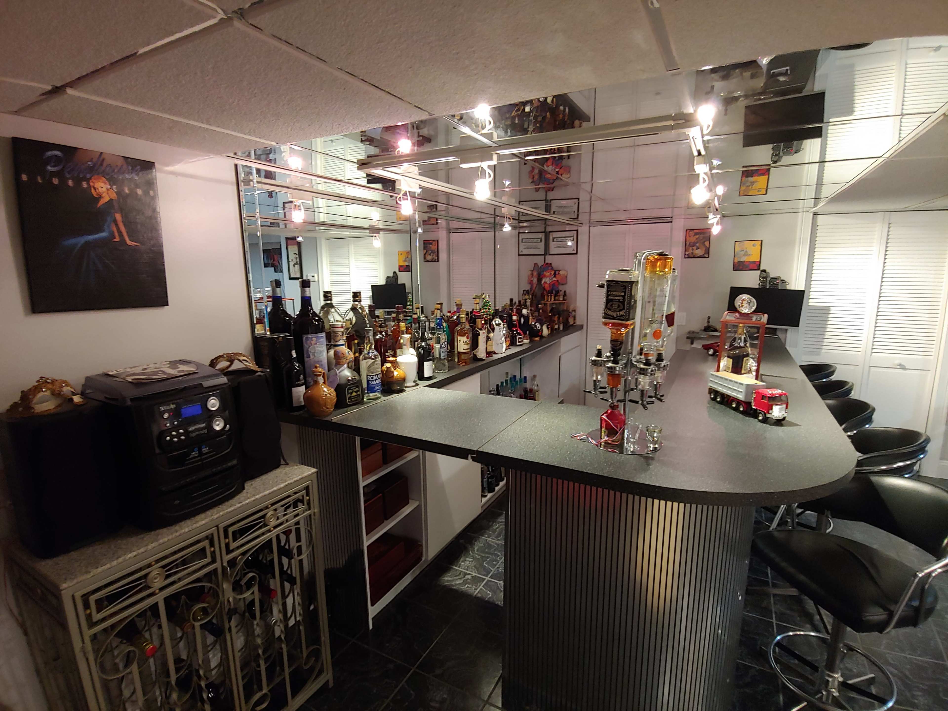 The image shows a well-stocked home bar featuring a curved counter with various bottles of alcohol displayed on shelves, a sound system, and bar stools.