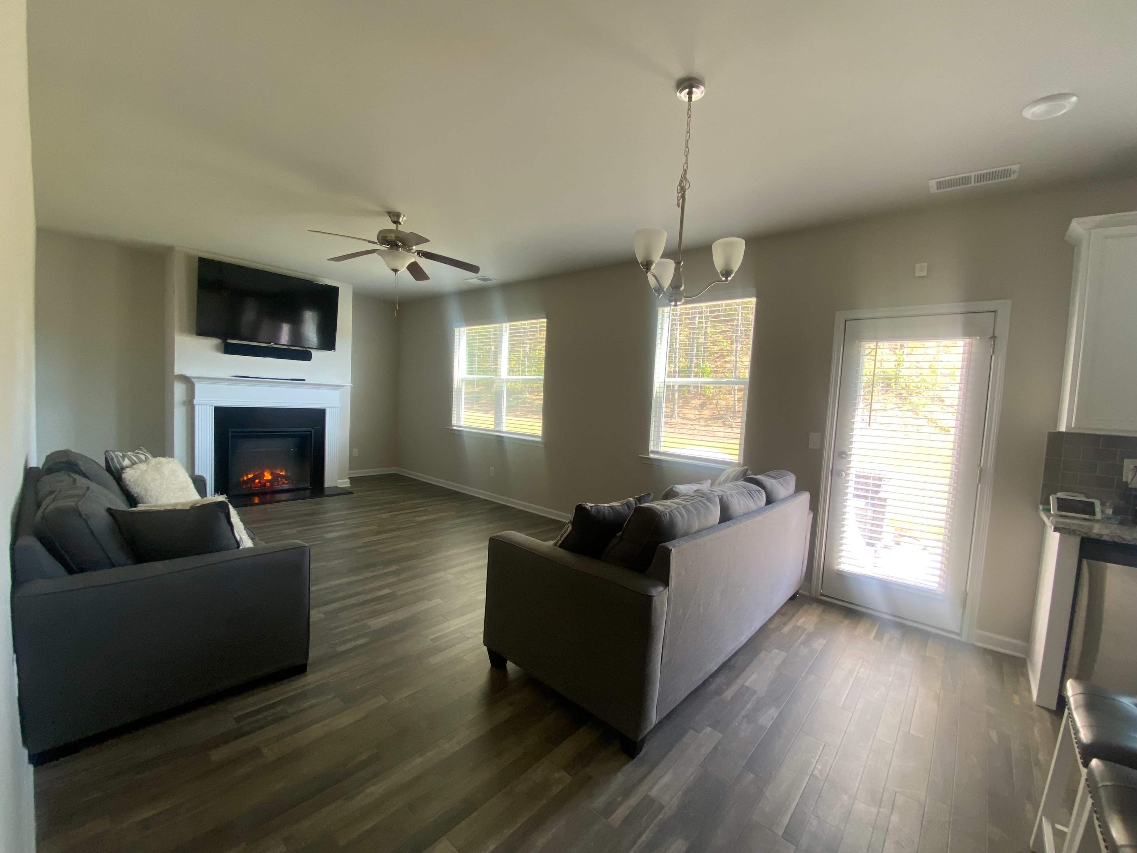A living room with a couch, a TV mounted above a fireplace, and a door leading outside, all illuminated by natural light from the windows.