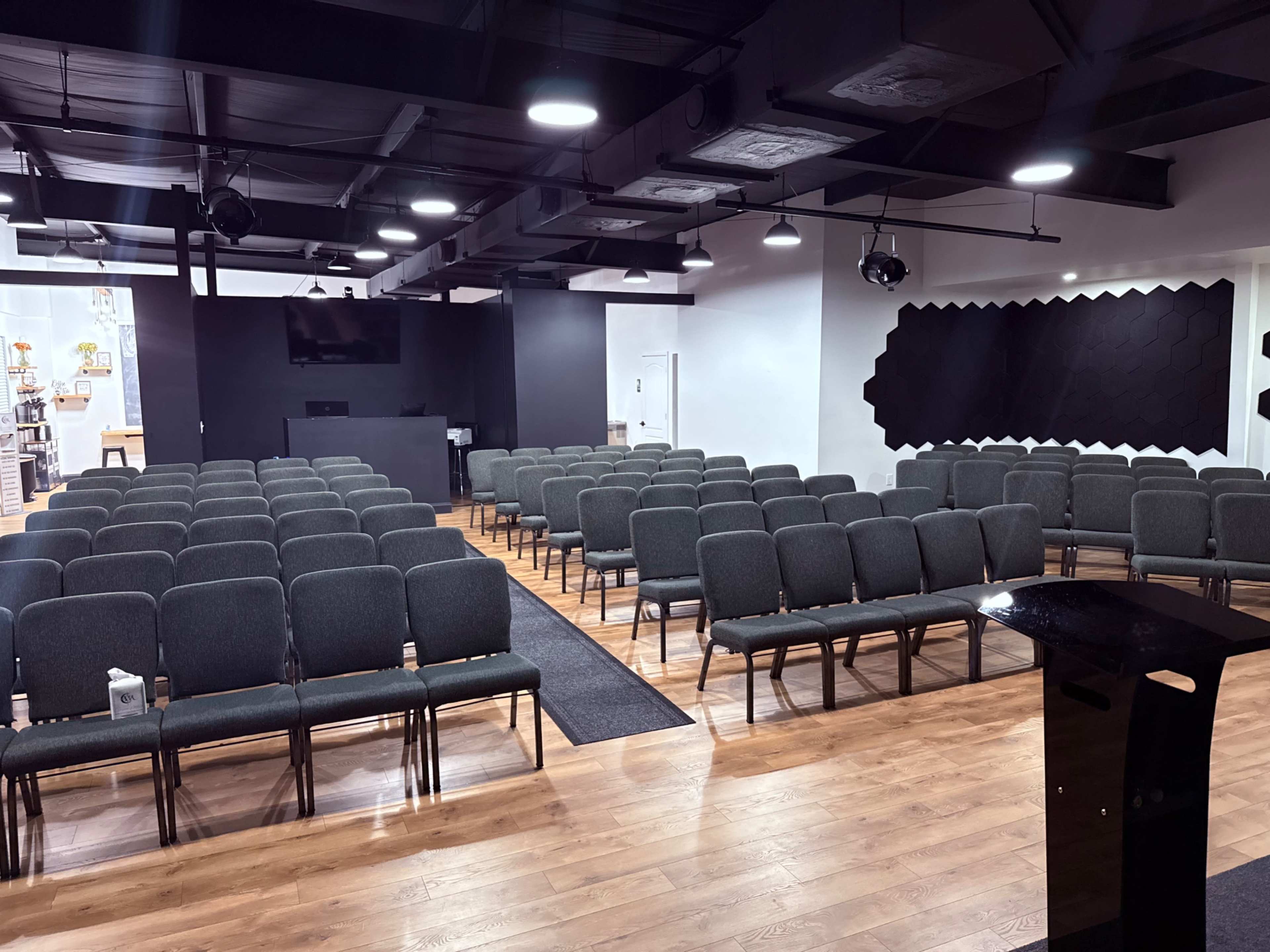The image shows a spacious room arranged with rows of gray chairs facing a black wall with a speaker system and a podium in front.