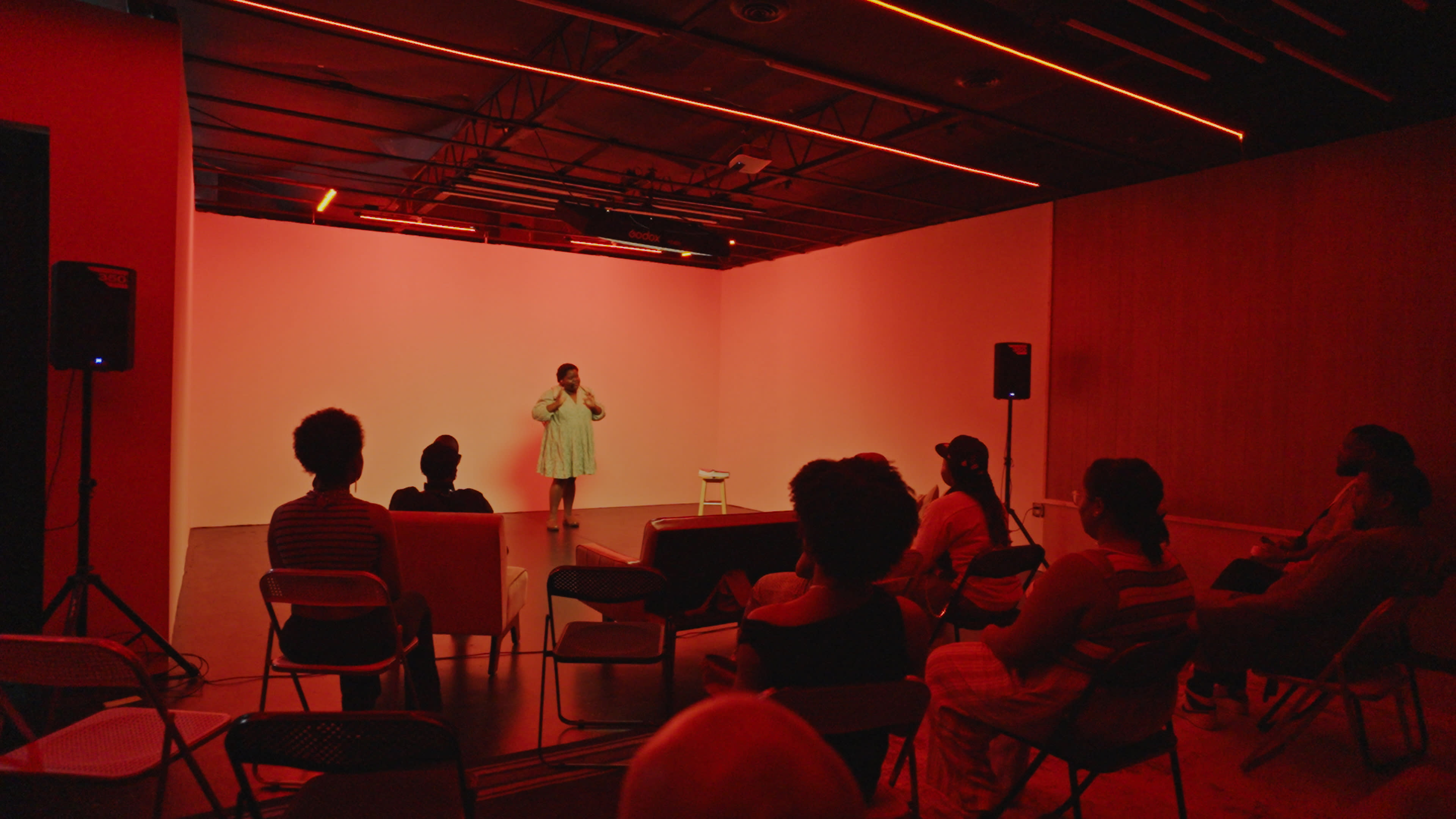 A performer stands on stage in a dimly lit room with red lighting while an audience watches from seated positions.