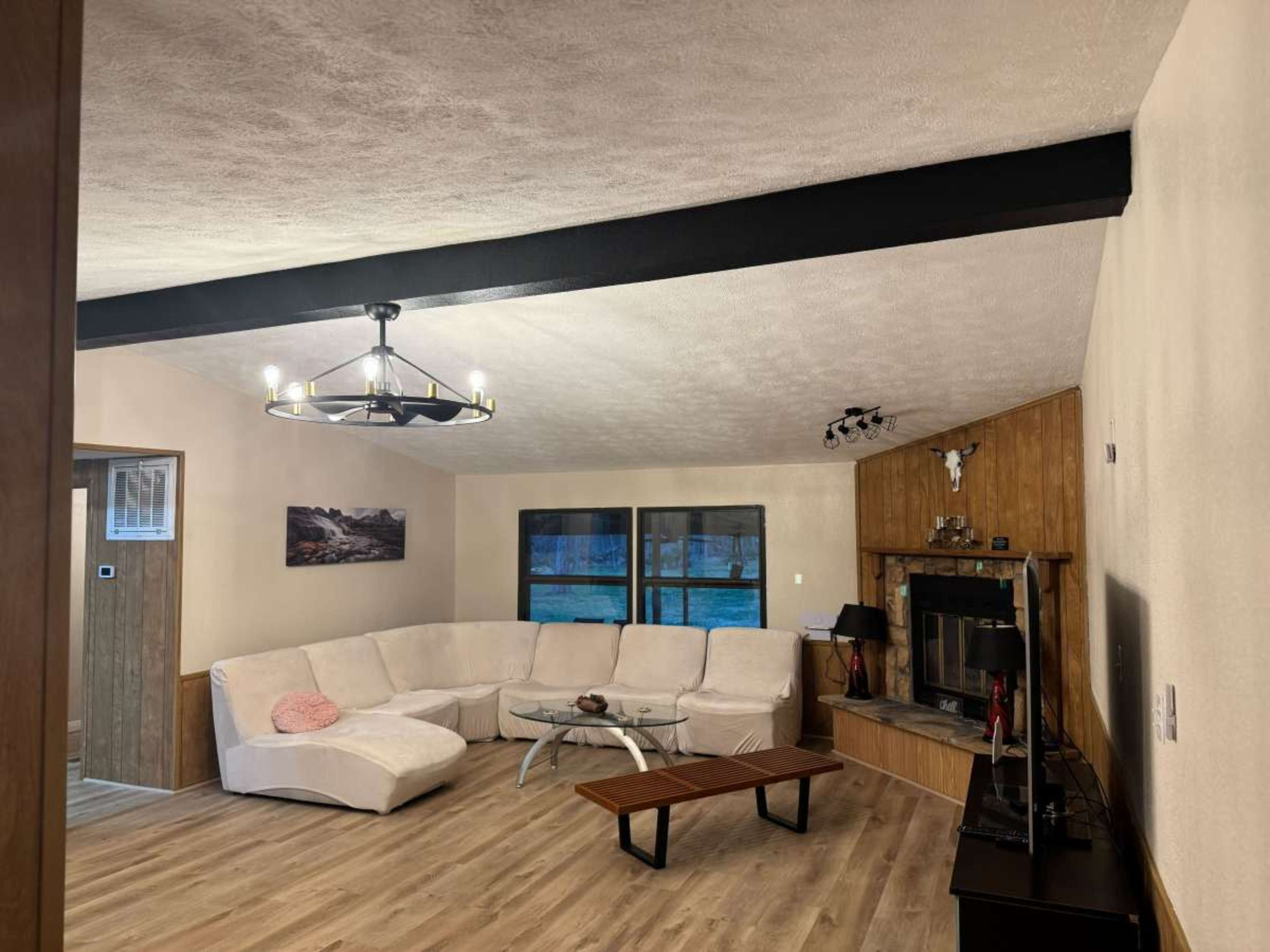 The image shows a spacious living room featuring a sectional sofa, a wooden coffee table, a fireplace with a wooden mantle, and a television on a black stand.