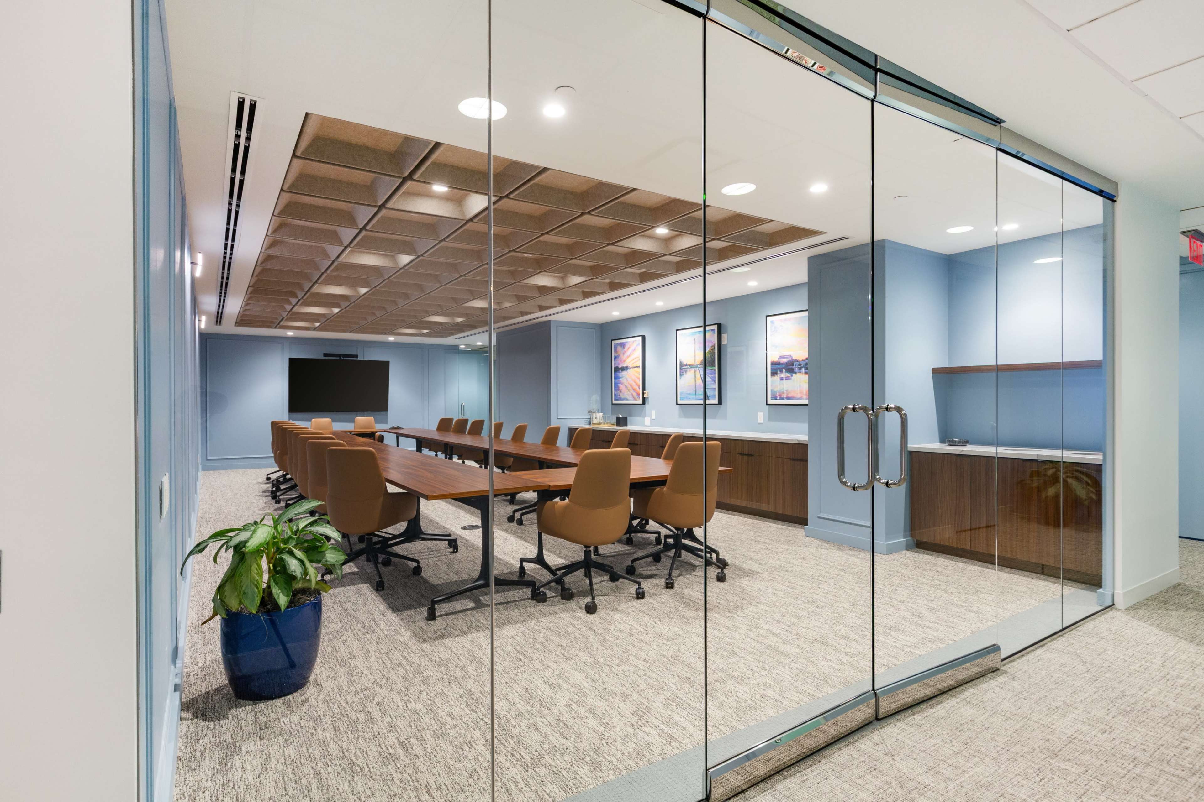The image shows a modern conference room with a large table, ergonomic chairs, and wall-mounted artwork, enclosed by glass walls.