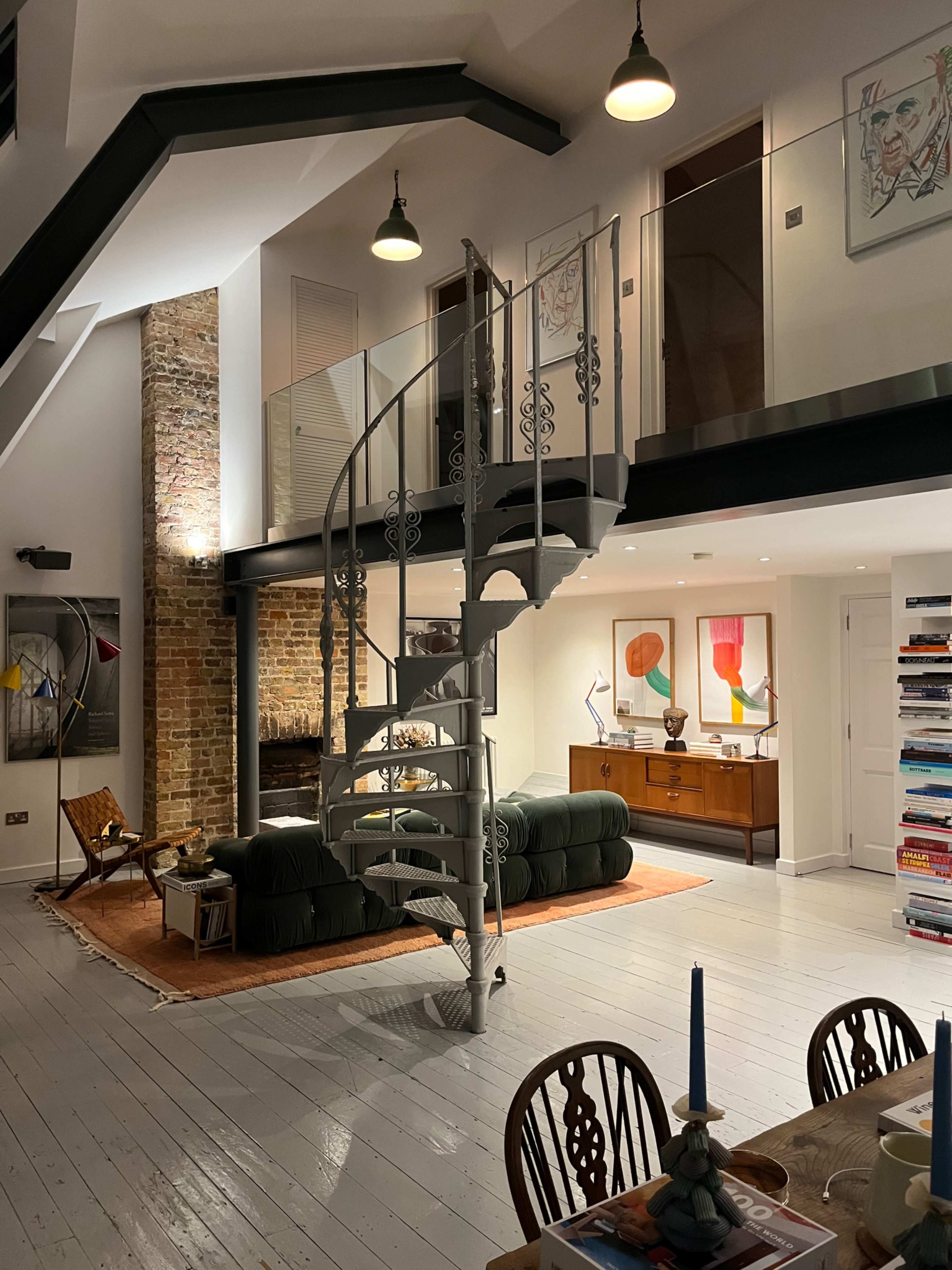 The image shows a modern loft interior featuring a spiral staircase, a comfortable seating area with artwork on the walls, and a dining table set with blue candles.