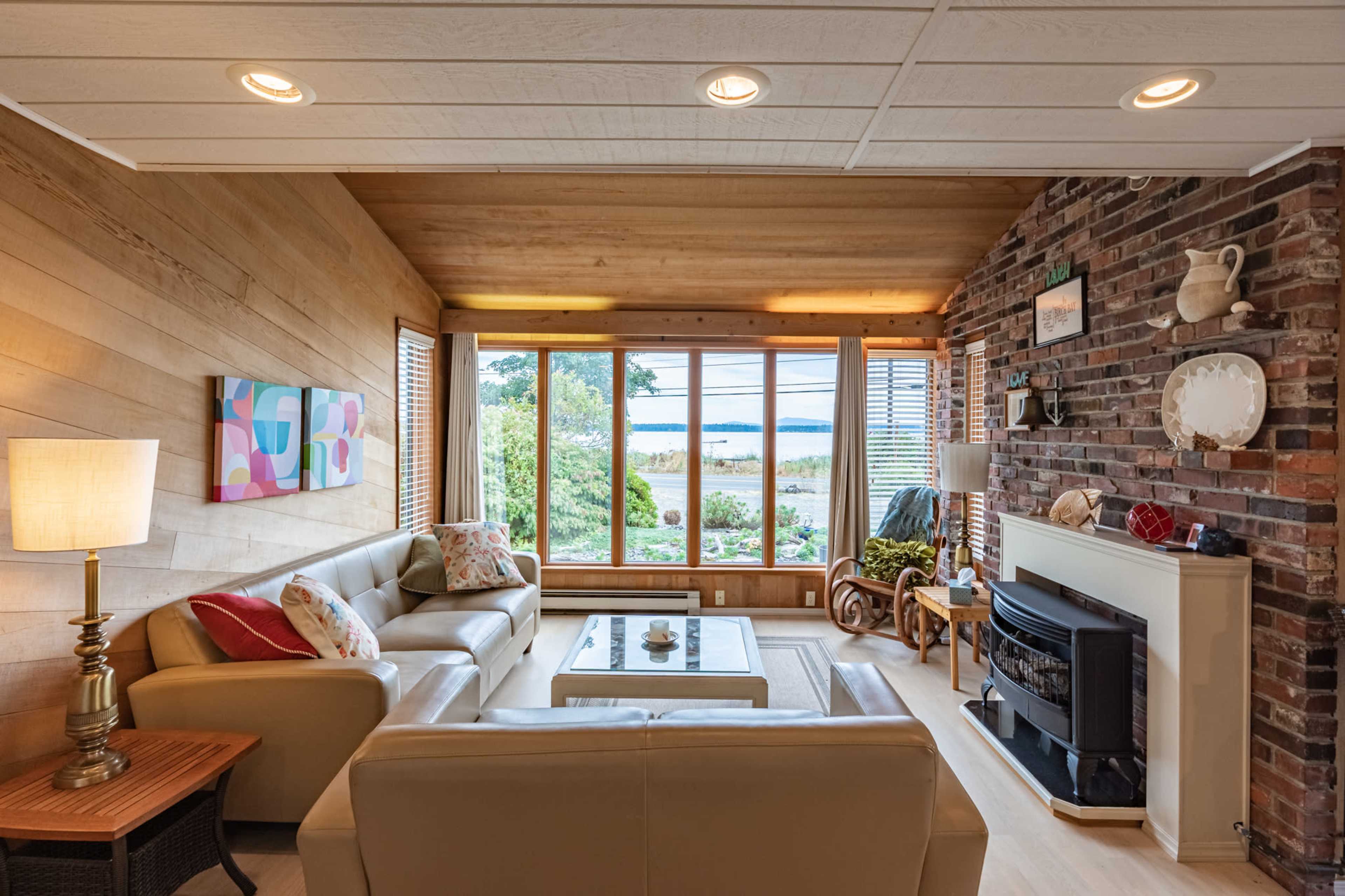 The living room features a beige sofa set, a lamp, and a fireplace, with large windows offering a view of the garden and water beyond.
