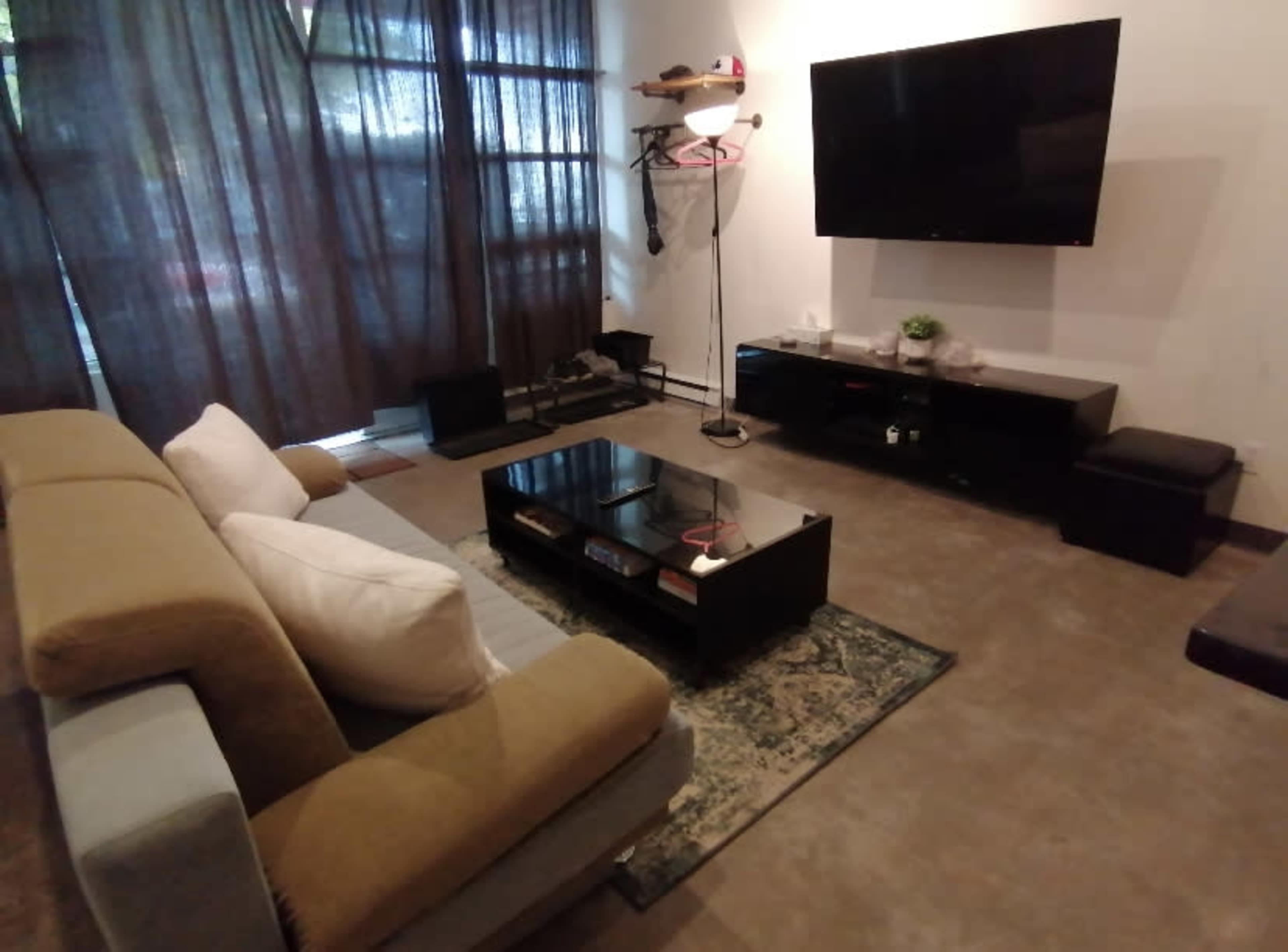 A living room features a couch with white and gray cushions, a dark coffee table, and a wall-mounted television, all set against a backdrop of sheer curtains.