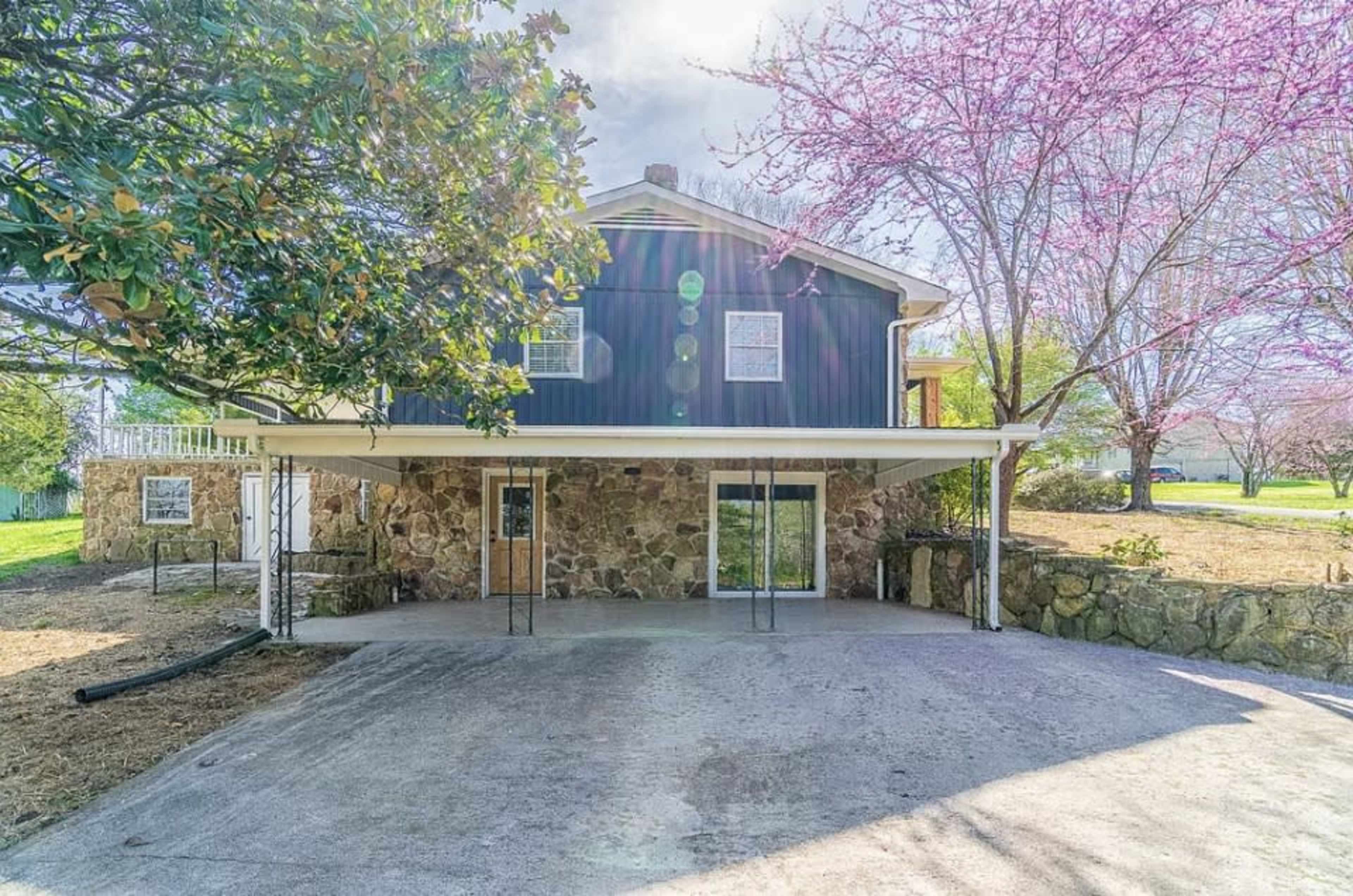 A two-story house with a stone facade and a covered patio is situated in a grassy area with blooming trees.
