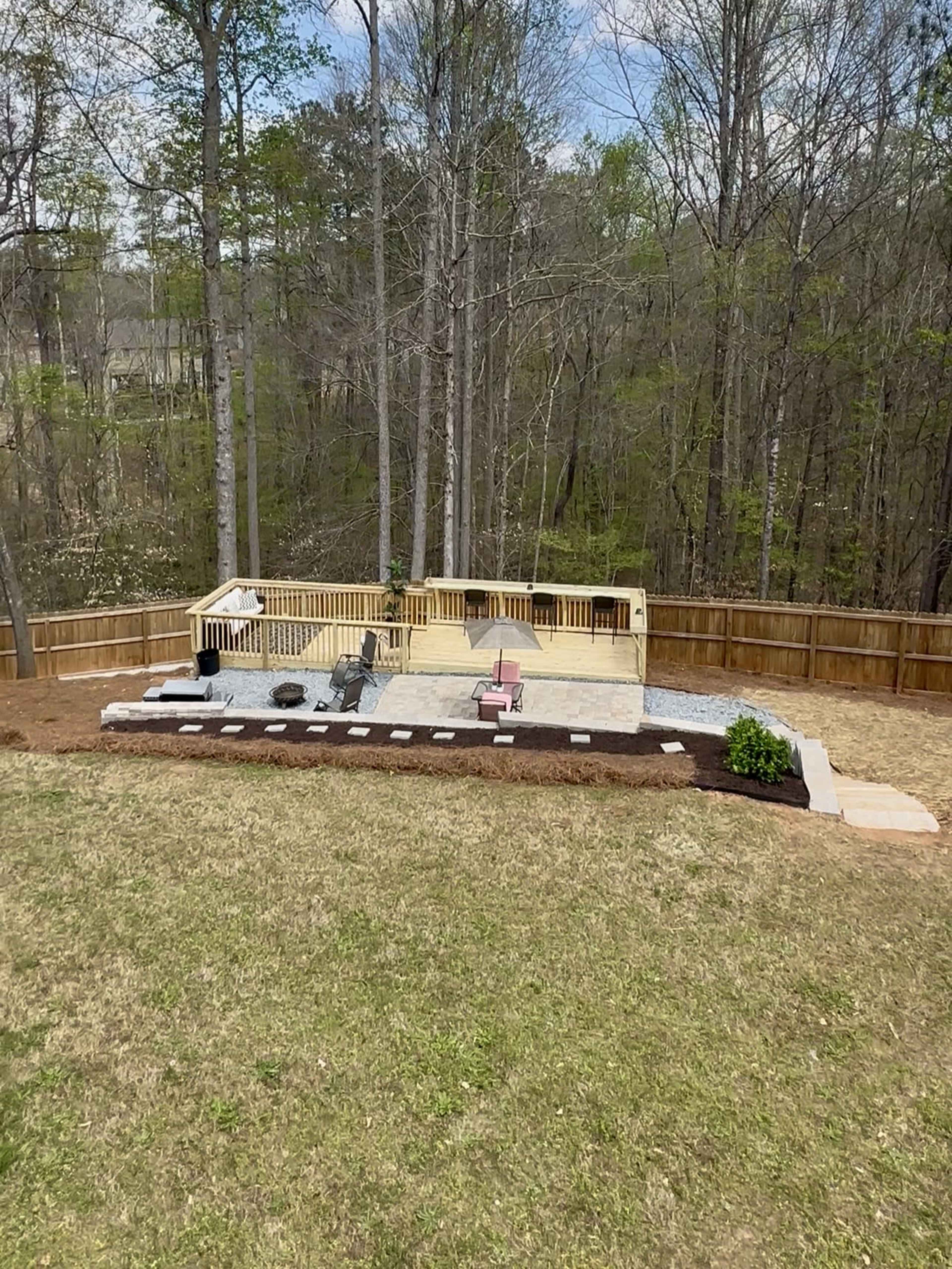 The image shows a wooden deck area with two chairs and a railing, surrounded by a landscaped yard featuring a stone path, mulch beds, and trees in the background.