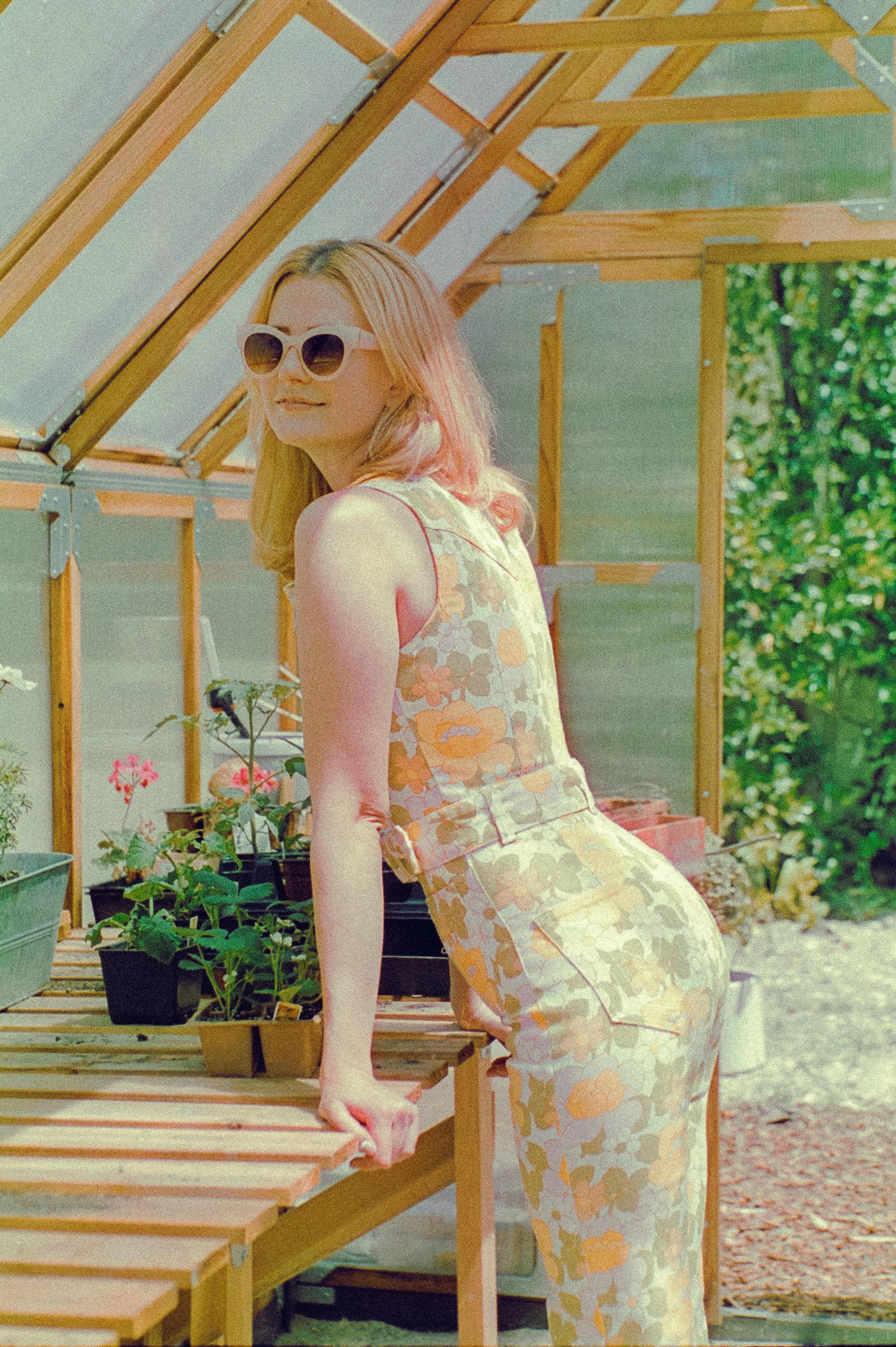 A woman in sunglasses poses next to a table full of potted plants in a greenhouse.