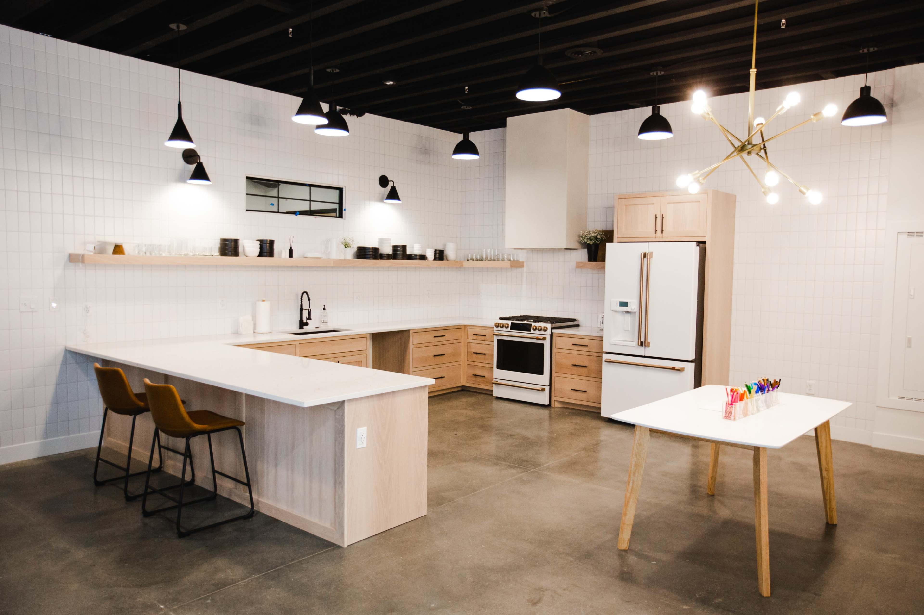 A modern kitchen with a central island, a refrigerator, an oven, and a dining table surrounded by chairs, all set against a backdrop of white tiled walls.