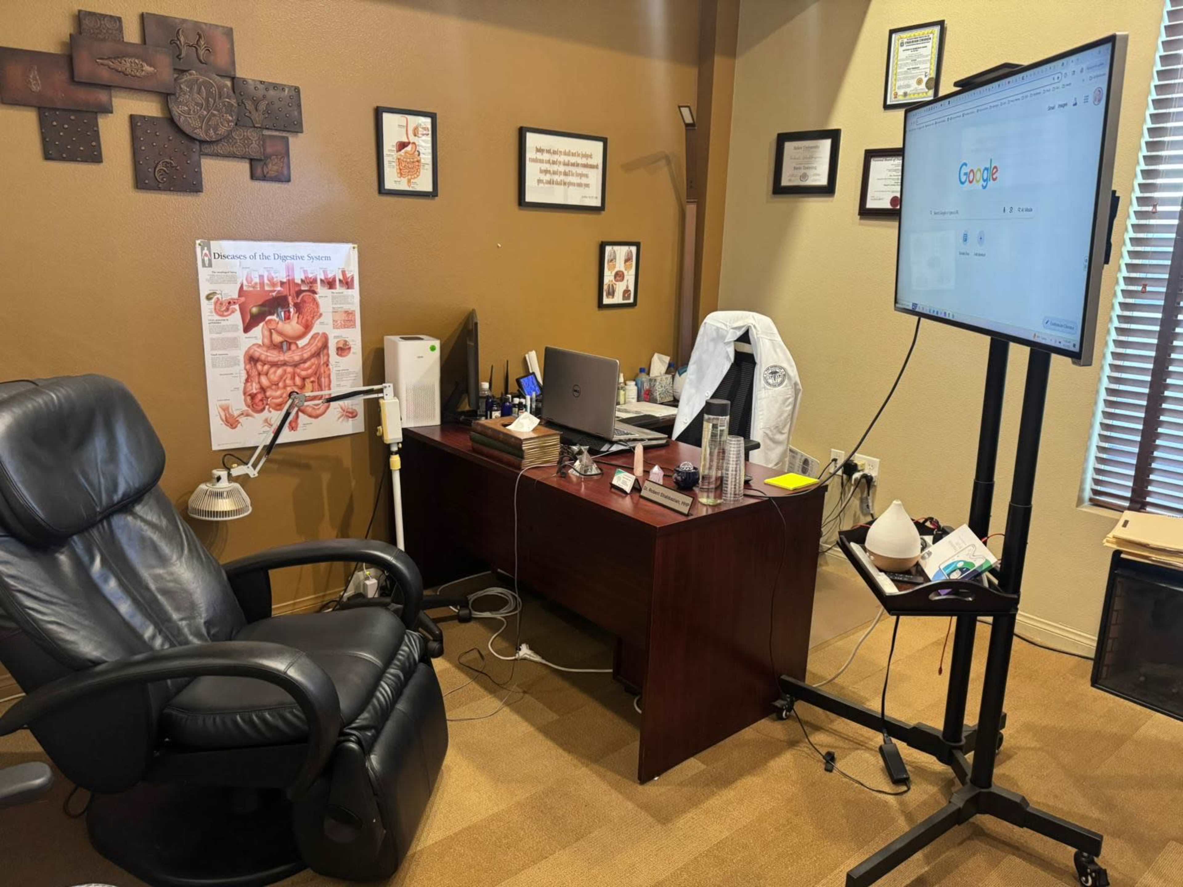 The image shows an office space featuring a leather recliner, a wooden desk with a computer and various items, a poster on the wall about the digestive system, and a large screen displaying the Google homepage.