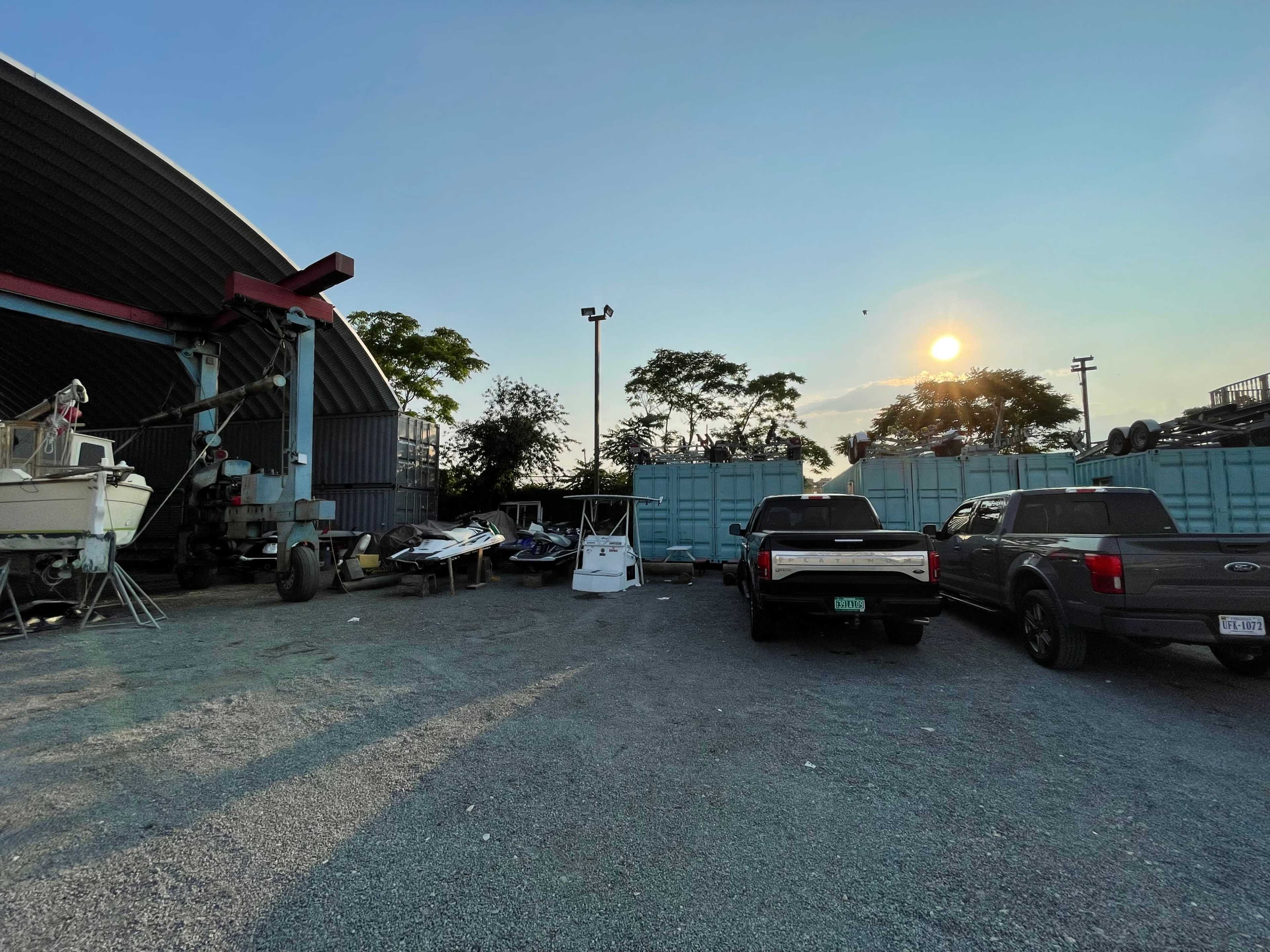 The image shows a storage area with several trucks and a boat, surrounded by shipping containers under a sunset sky.