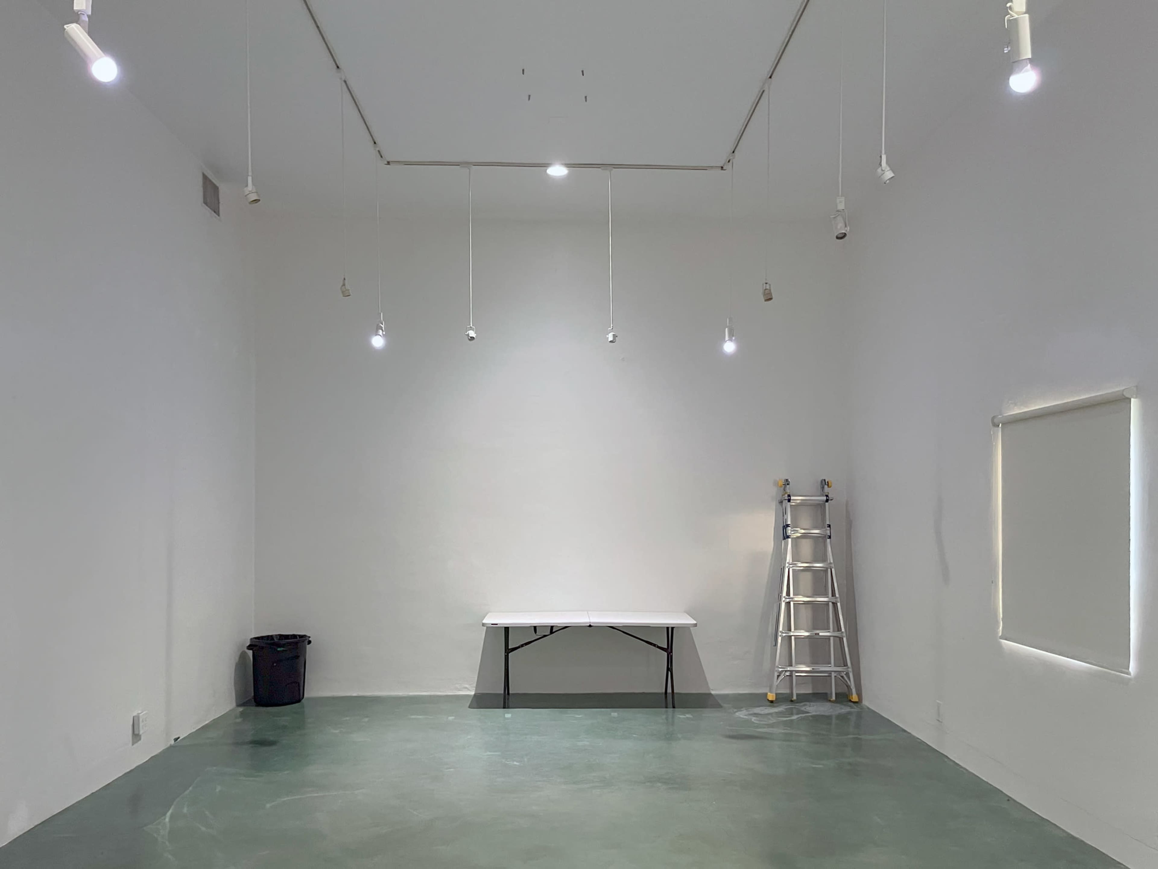An empty room with a green floor, a white wall, a foldable table in the center, a ladder against the wall, and a trash bin in one corner.