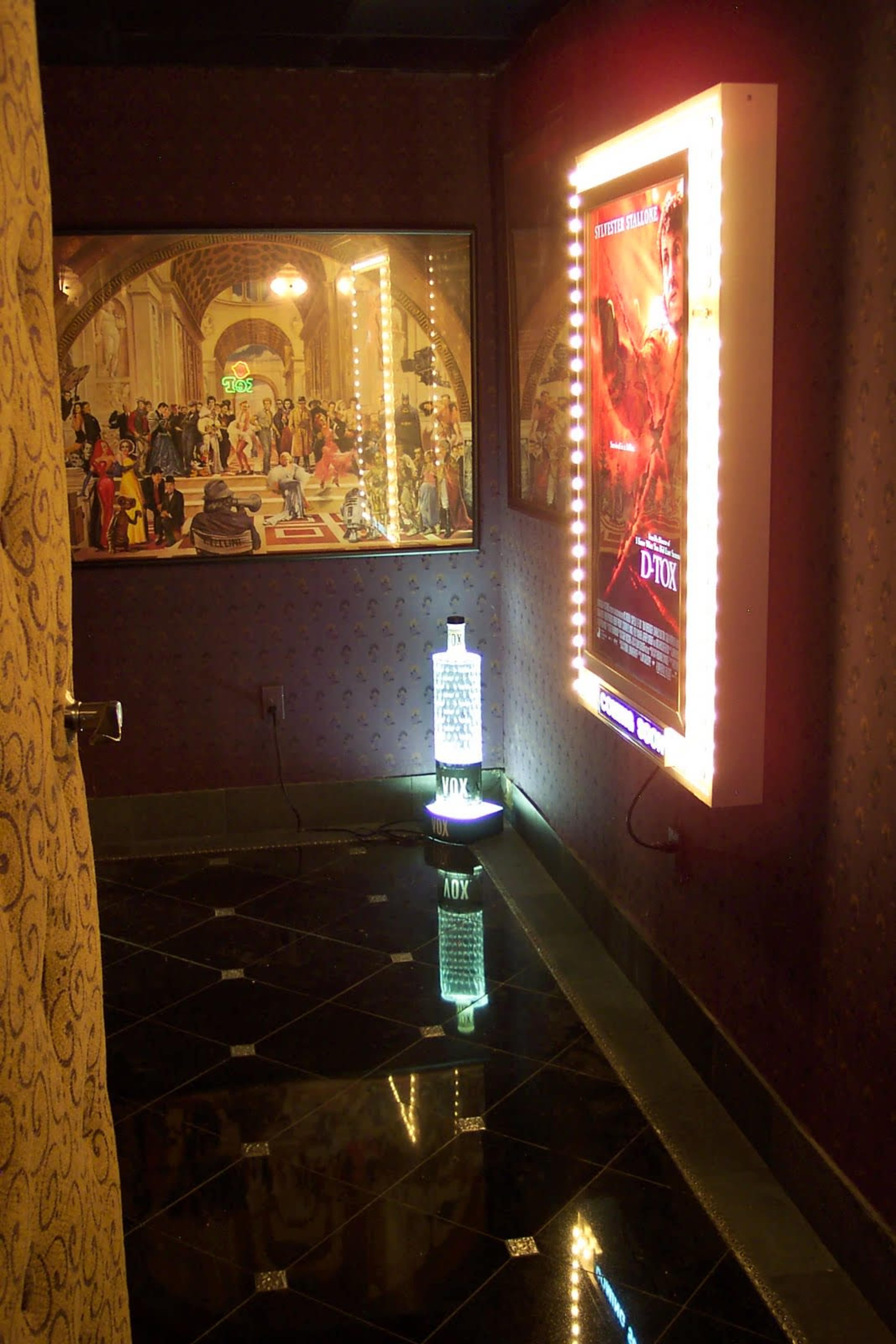 A dimly lit corridor with ornate wall decor, featuring a brightly lit movie poster and a decorative display illuminating a bottle on the floor.