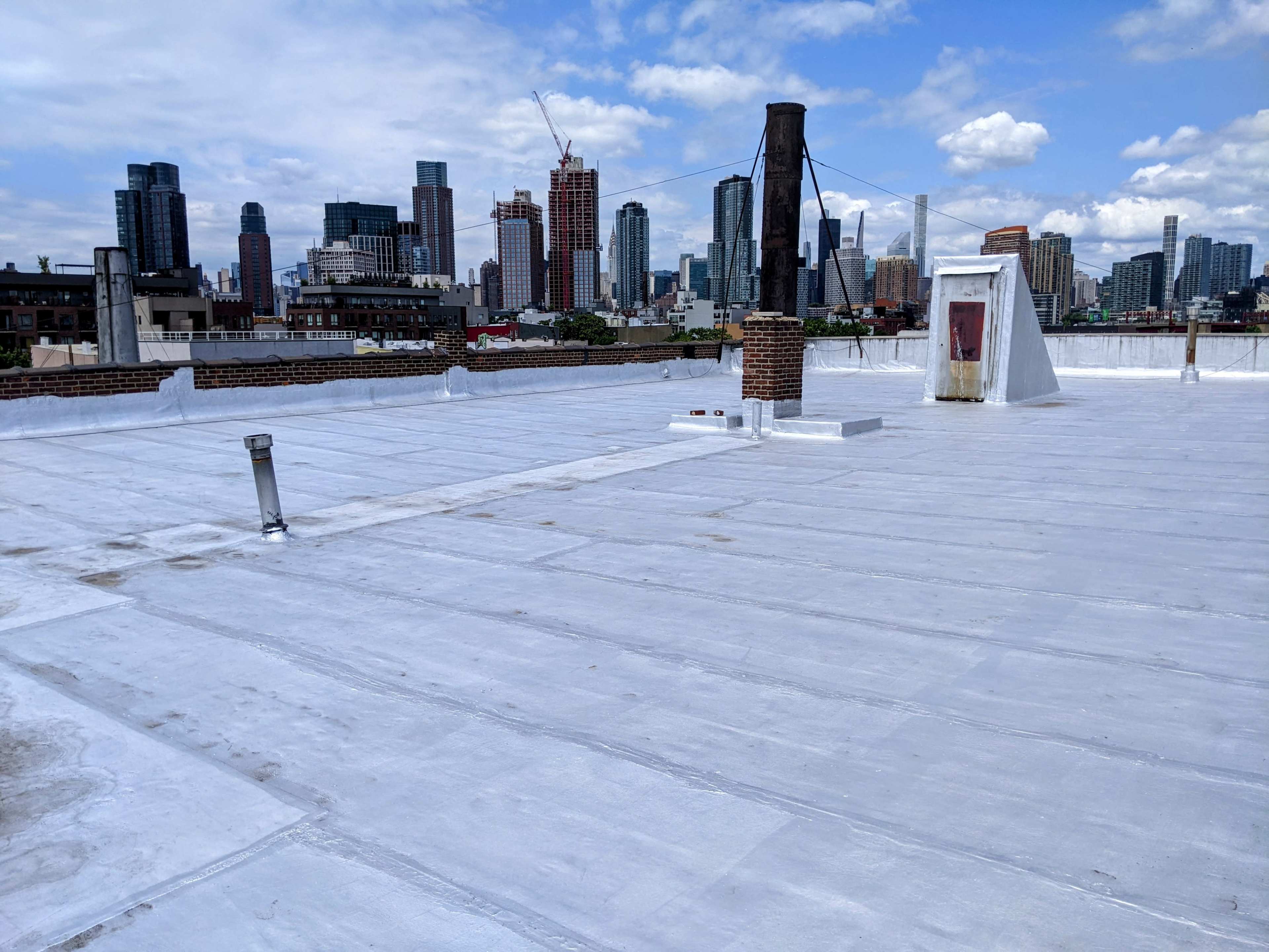 A flat rooftop with a white surface overlooks a city skyline featuring various tall buildings under a partly cloudy sky.