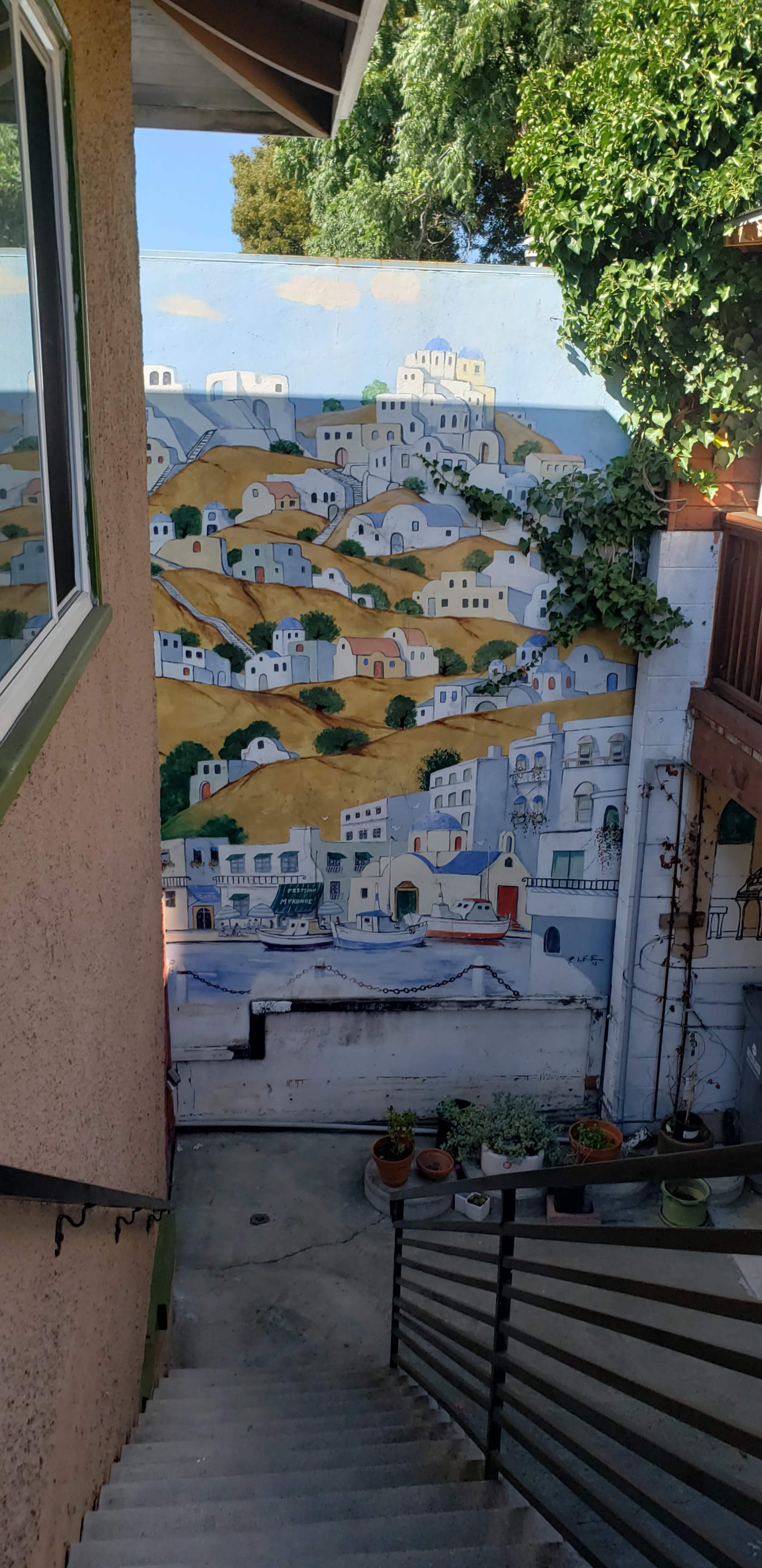 The image shows a mural depicting a hillside village with white buildings and vibrant greenery, located in a courtyard surrounded by stairs and potted plants.