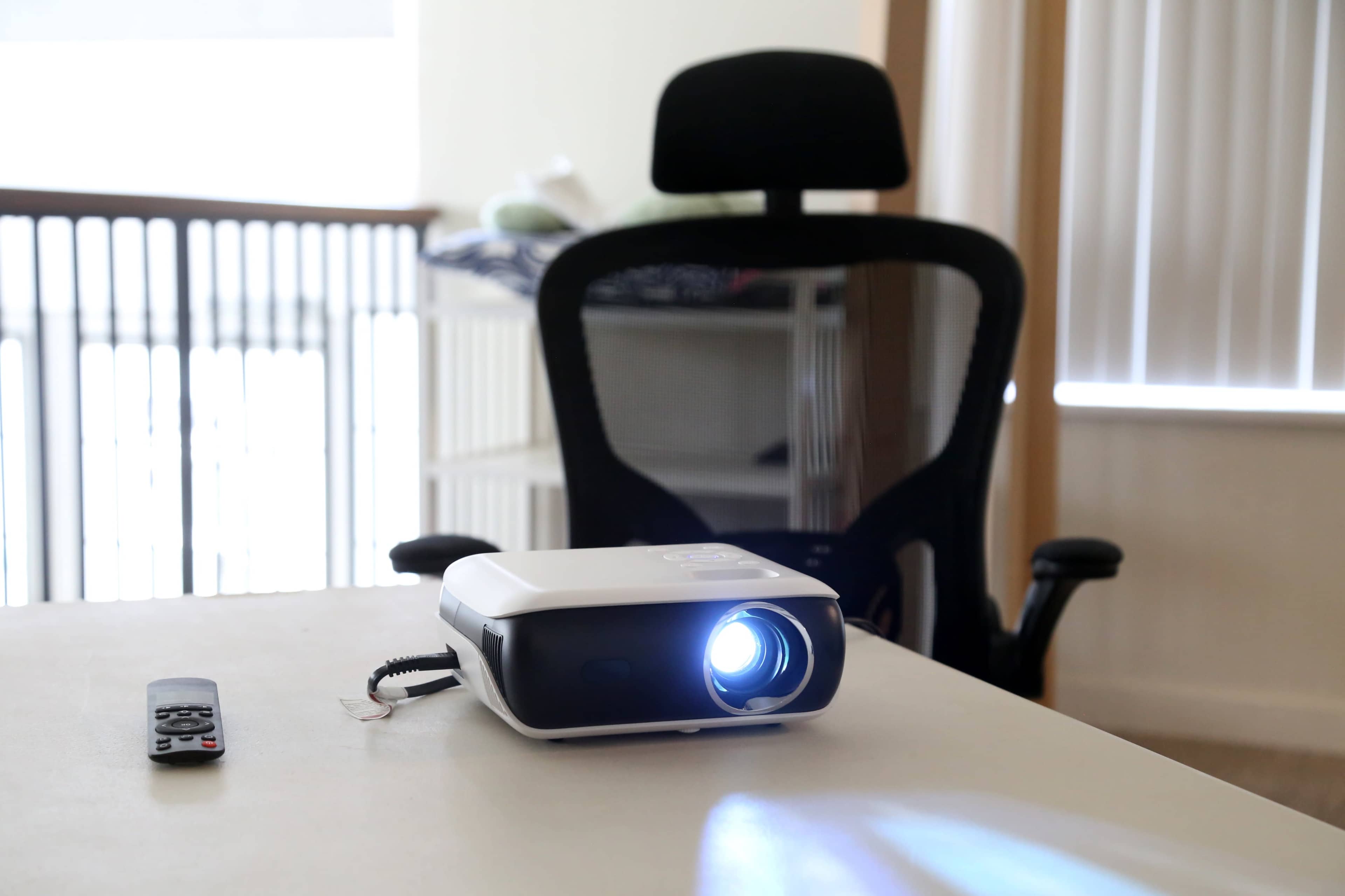 A projector is positioned on a table with a remote control nearby, while a black office chair is visible in the background.