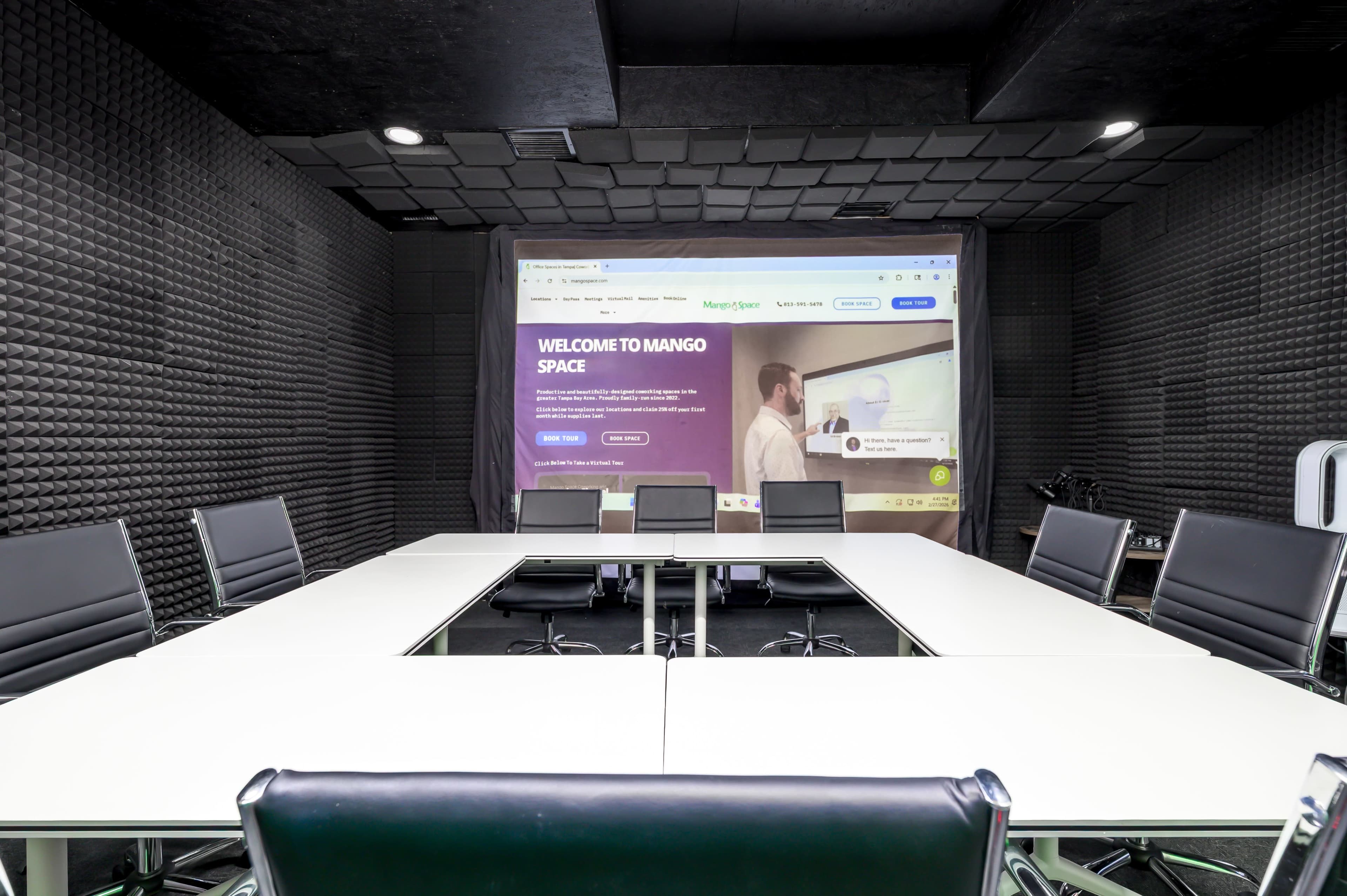 A modern conference room features a large table surrounded by chairs, with a screen displaying a welcome message on a dark textured wall.