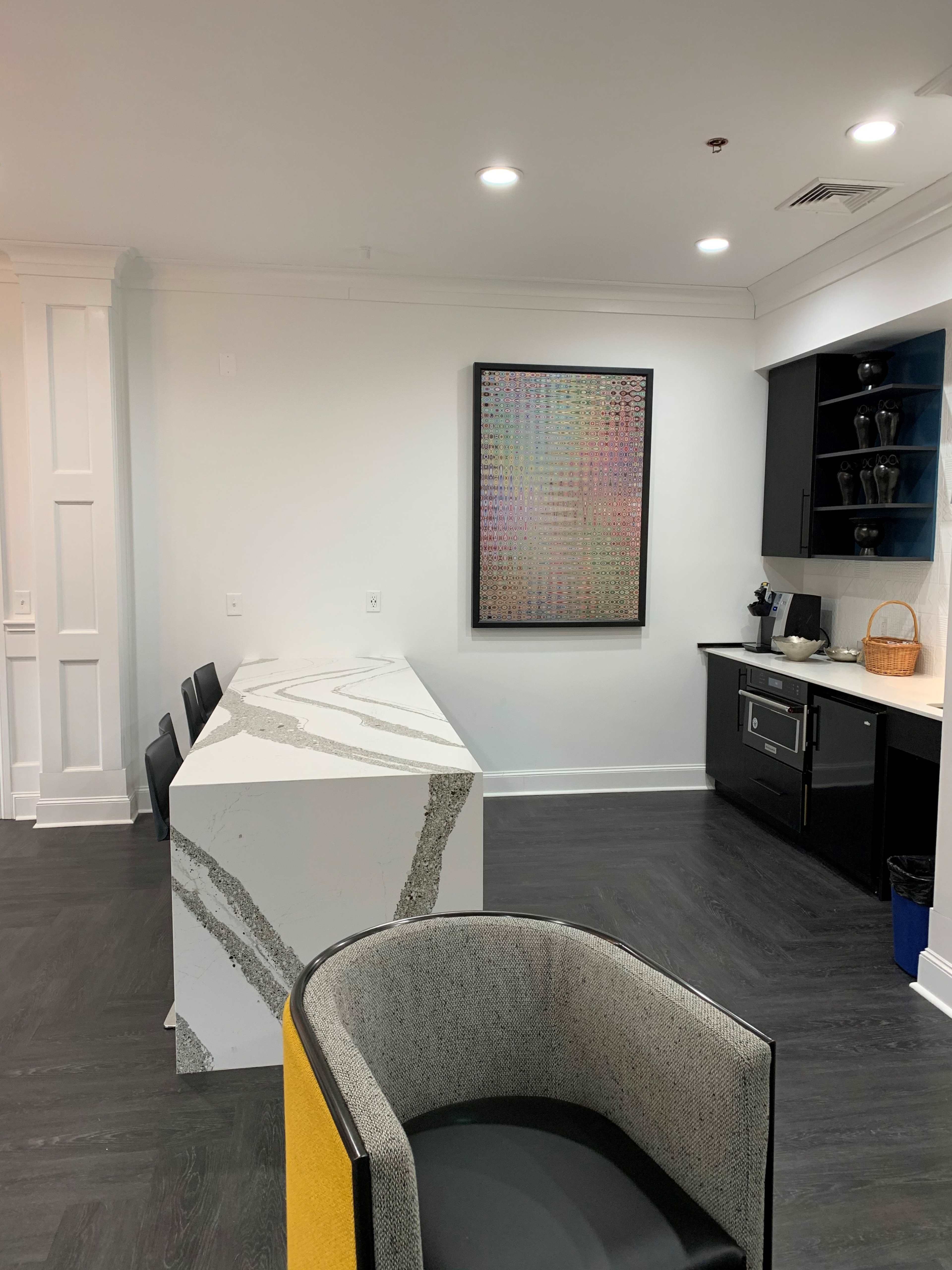 The image shows a modern kitchen area with a large, white marble island, black cabinetry, and a colorful piece of artwork hanging on the wall.