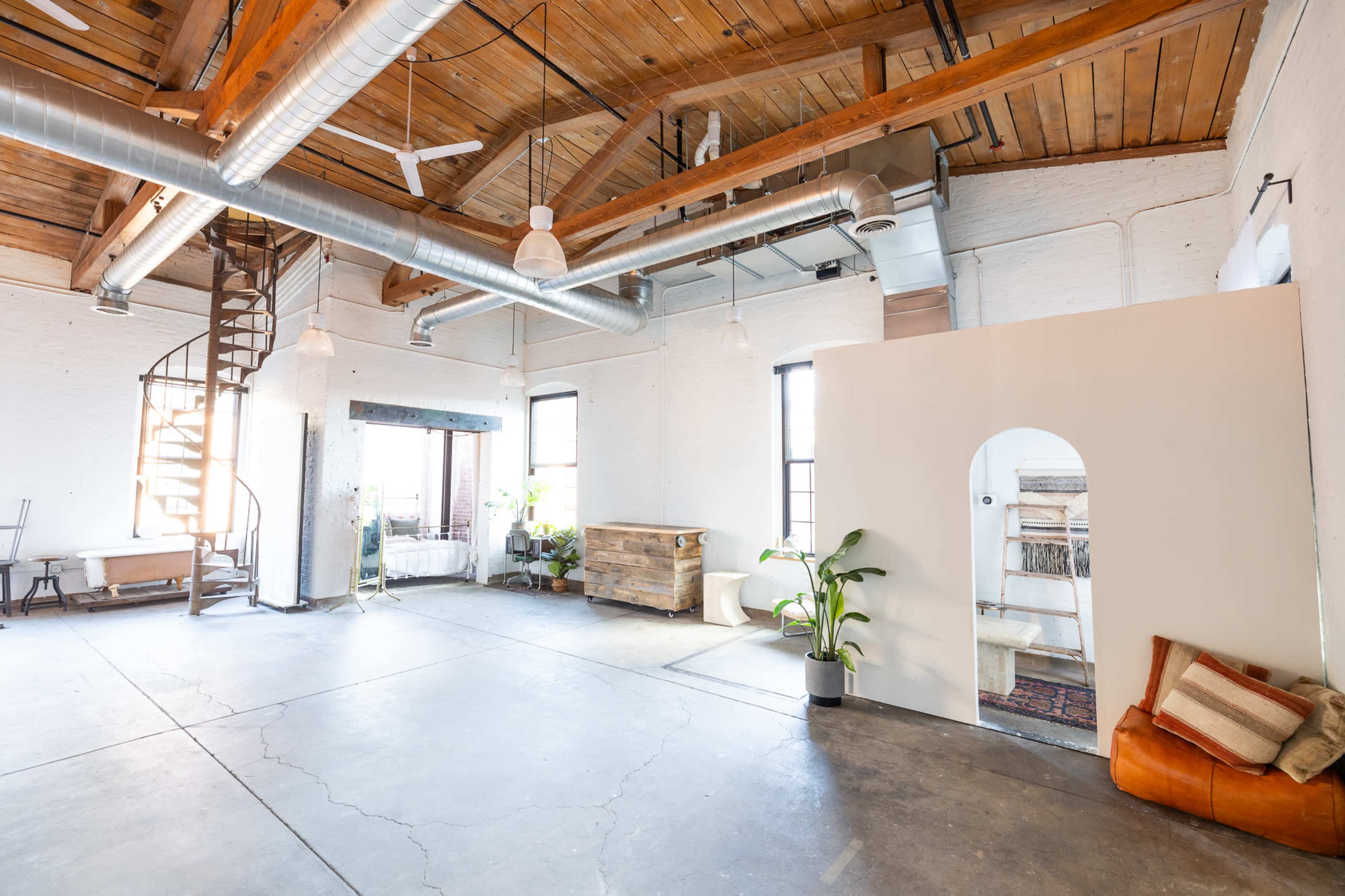 A spacious, modern loft with high wooden ceilings, exposed ductwork, and a mix of industrial and cozy furnishings, including a spiral staircase and plants.