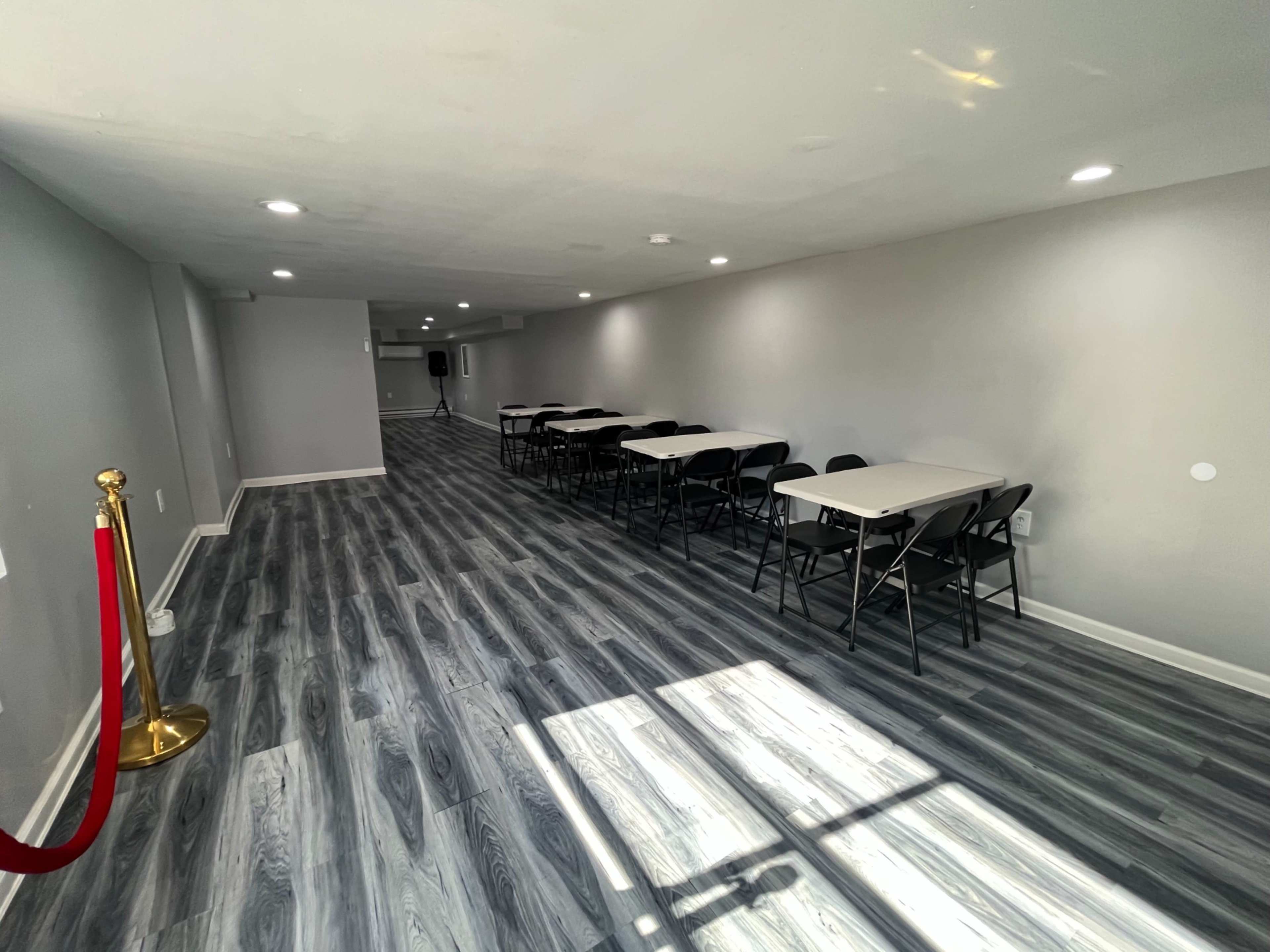 A spacious, well-lit room with several tables and chairs arranged in rows along the length of the floor, which has a gray wood-like finish.
