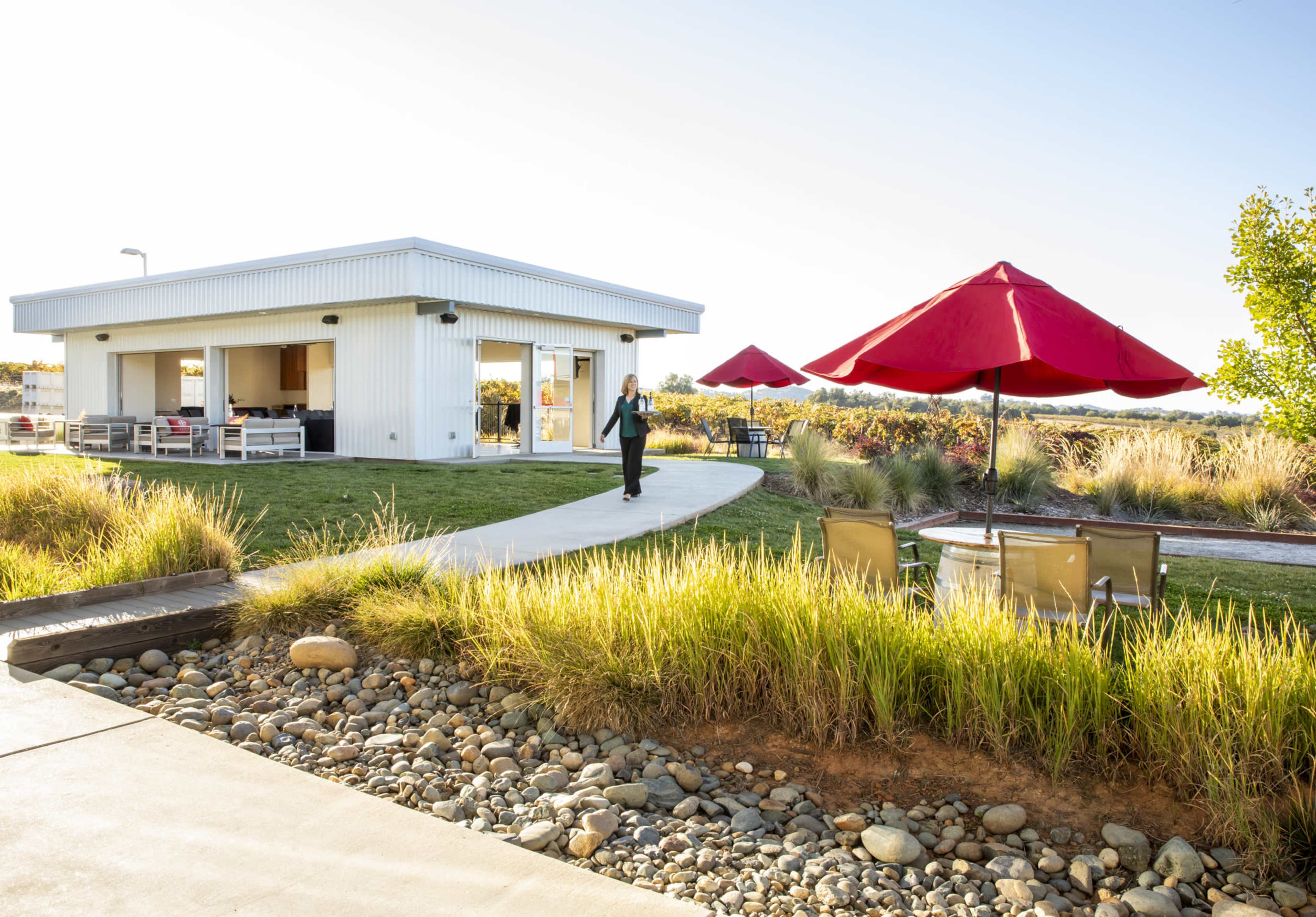 A woman walks along a path toward a white building surrounded by green grass and red umbrellas.