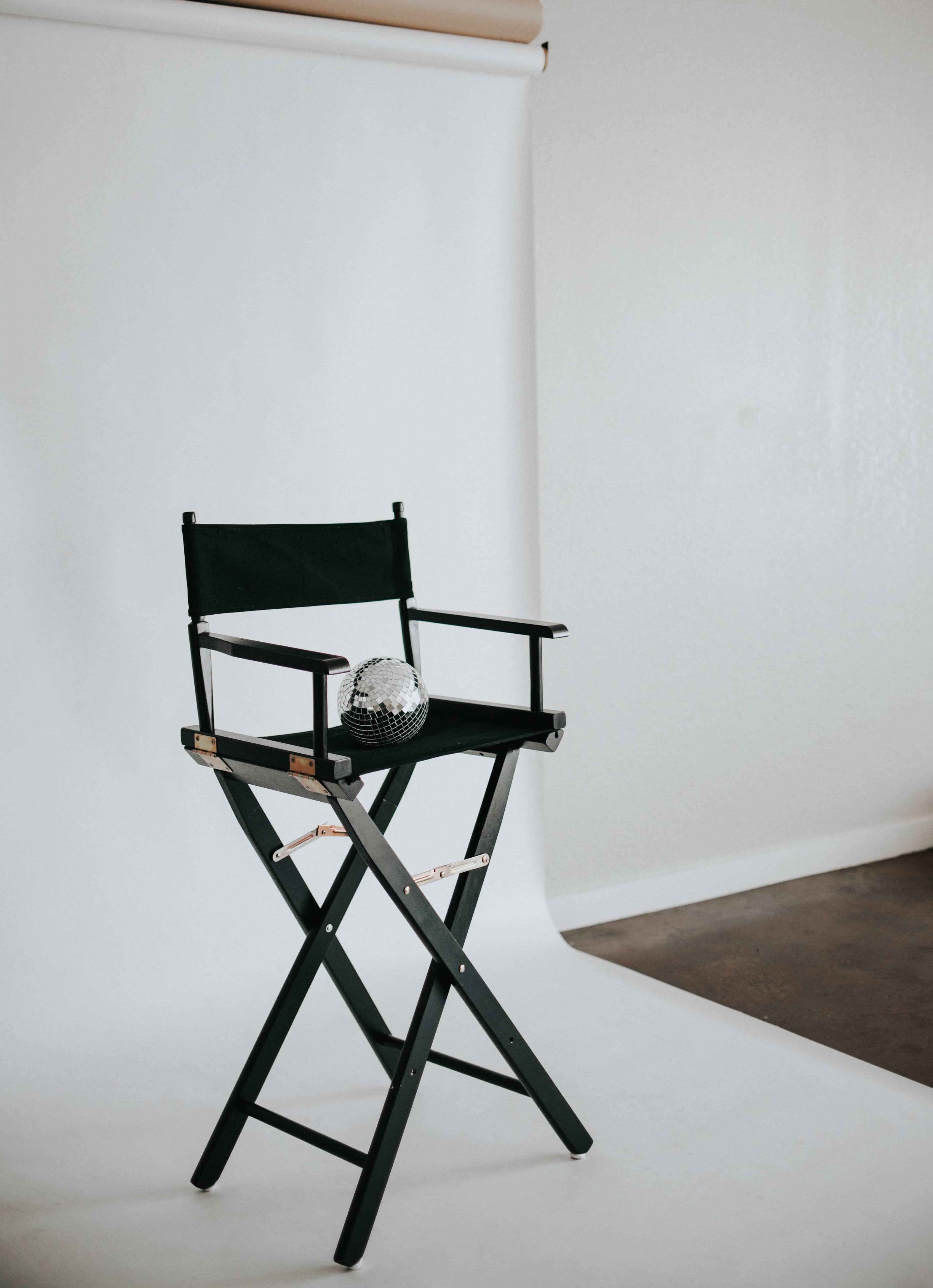 The image shows a black director's chair positioned in front of a plain white backdrop with a small disco ball placed on the seat.