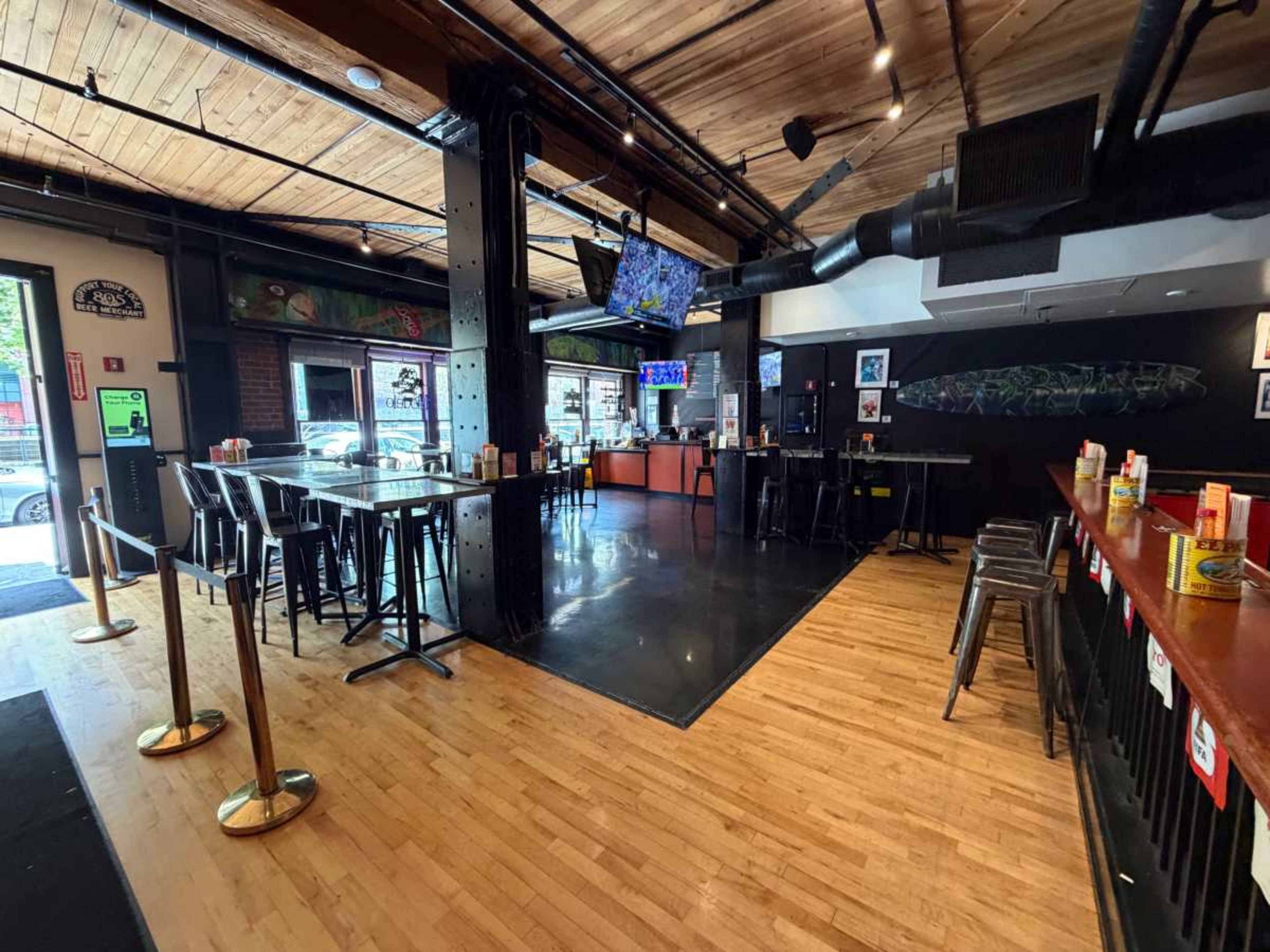 The image shows an interior view of a sports bar with high-top tables and a bar counter, featuring wooden floors and a large television mounted on the wall.