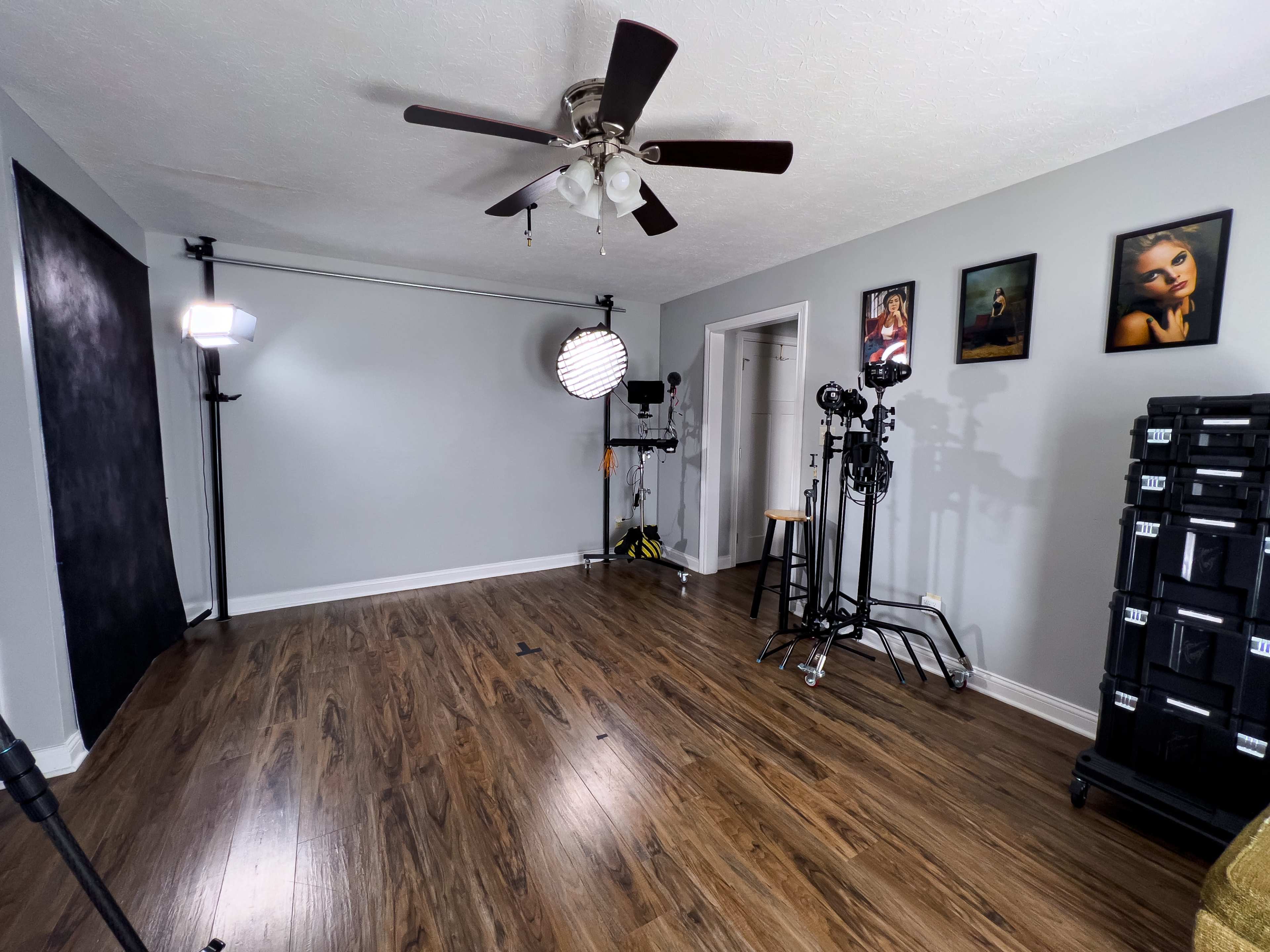 The image shows a spacious room with hardwood floors, featuring photography equipment, a ceiling fan, and several framed artworks on the walls.