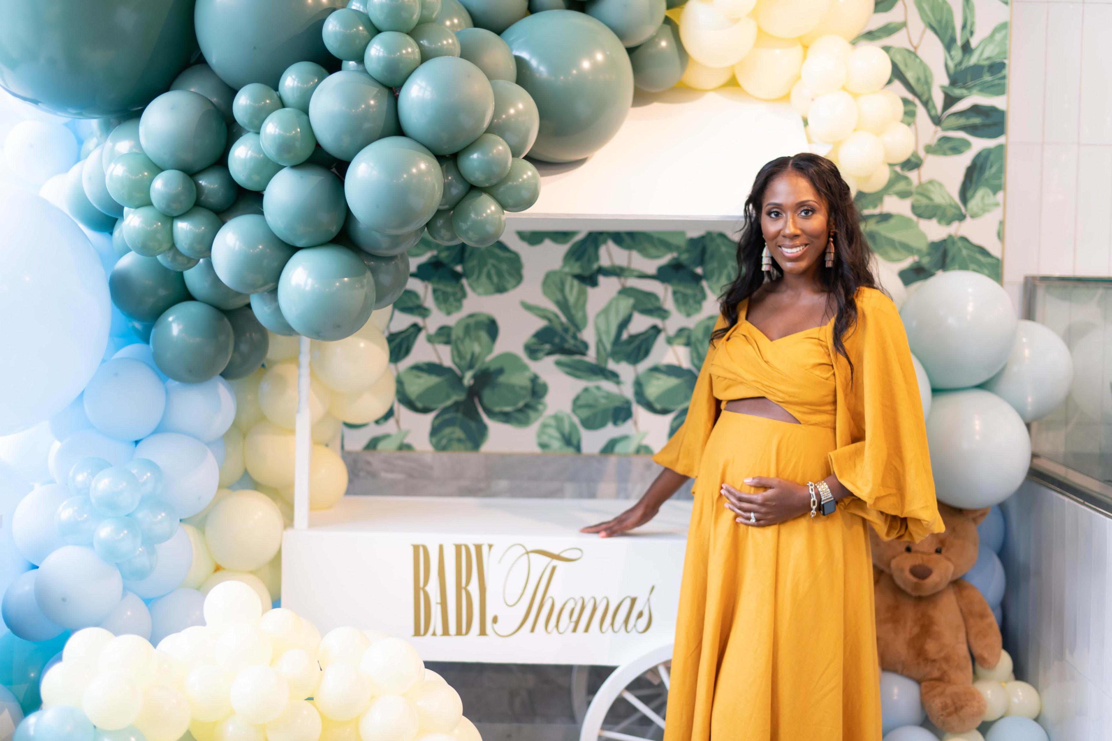 A woman in a yellow dress standing in front of balloons for a baby shower. 