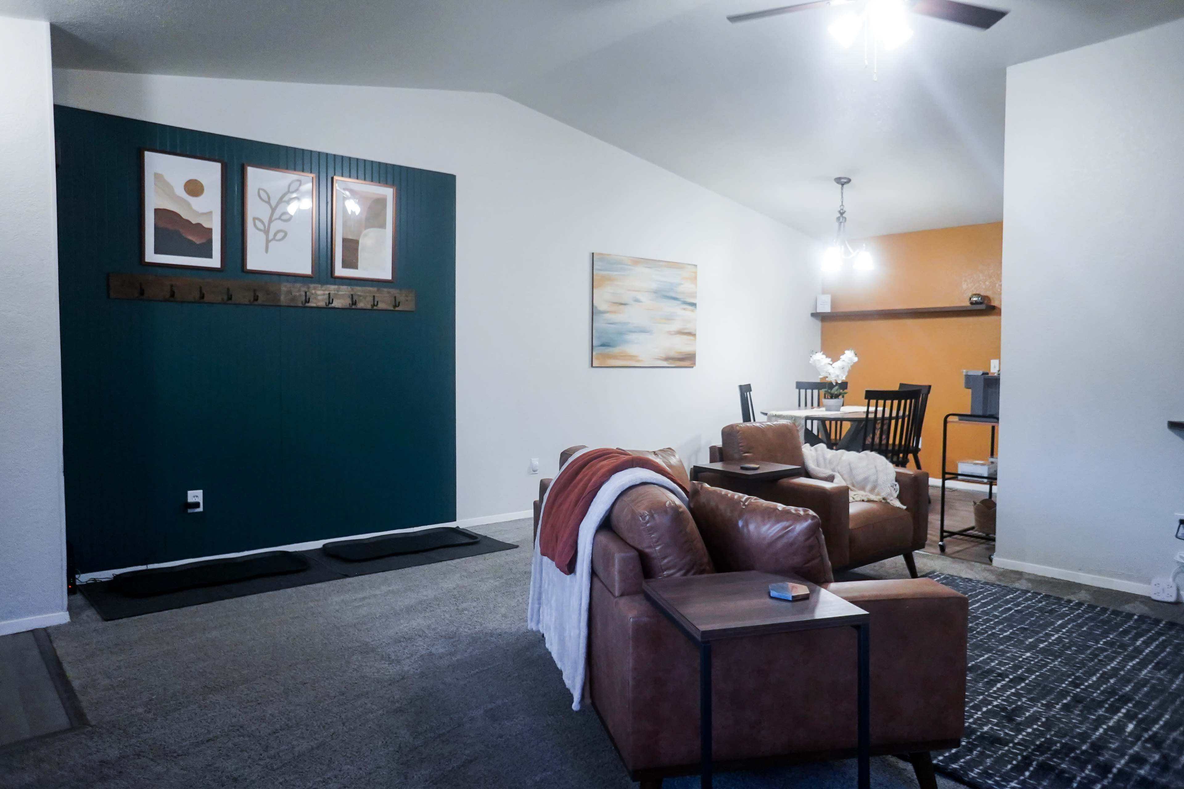 The image shows a cozy living room with a brown leather sofa, a small side table, and a dining area in the background featuring a wooden table and chairs.