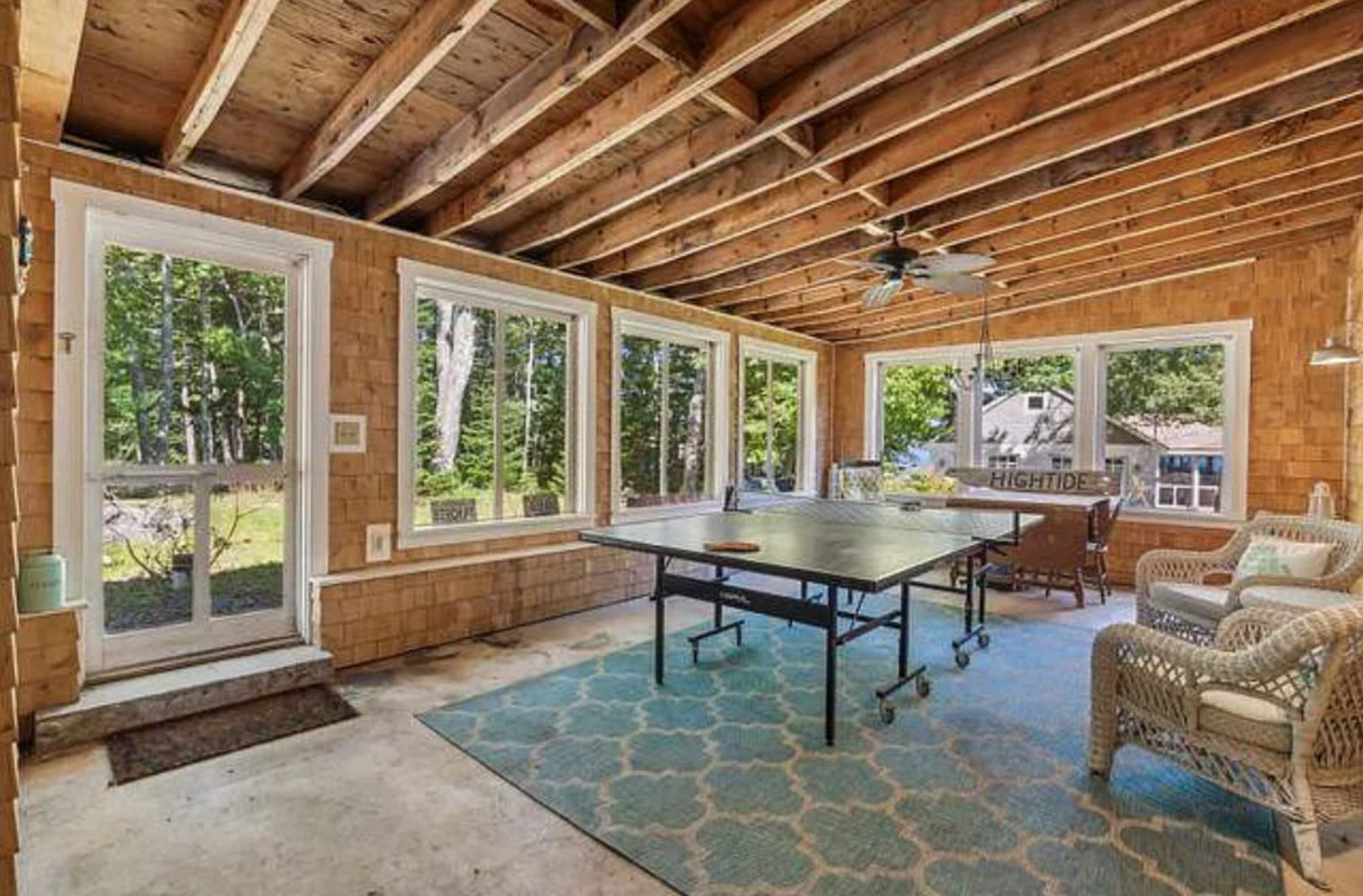 The image shows a partially completed sunroom with large windows, exposed wooden beams, and a ping pong table centered on a patterned rug.